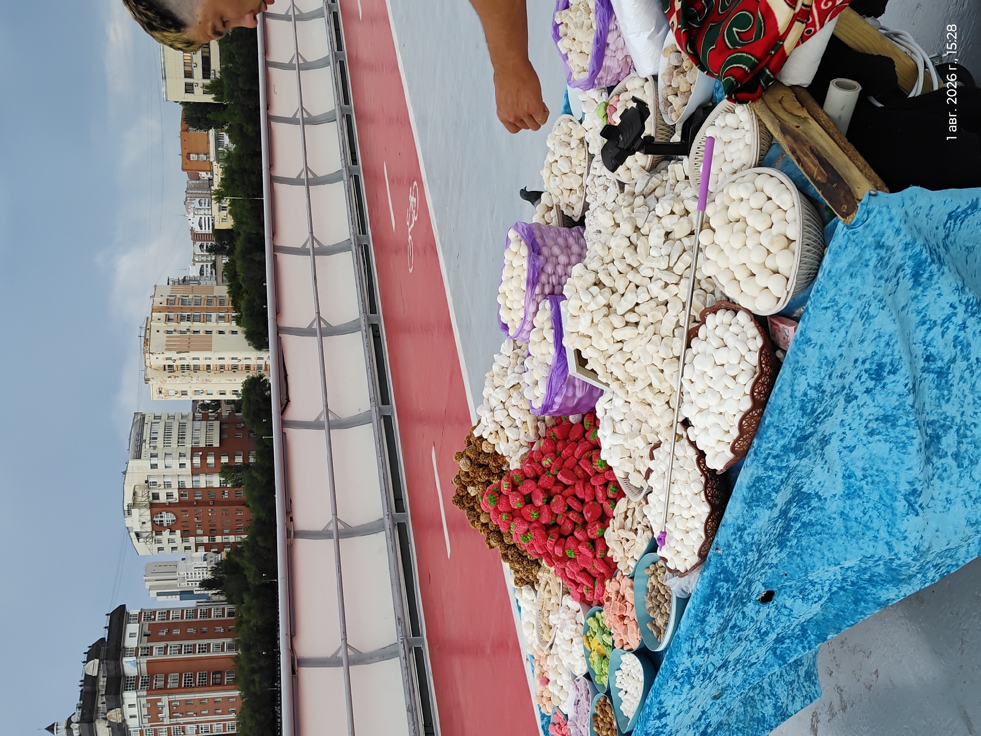 The image depicts an outdoor market stall laden with a variety of sweets, located on an elevated pedestrian walkway in Astana, Kazakhstan. In the background, several multi-story residential buildings with distinctive architectural styles are visible, suggesting an urban setting. The walkway itself is paved with gray material, and a red lane with a bicycle symbol indicates a designated path.
The stall is covered with a blue tarp and displays large piles of confectioneries. Prominently featured are red, strawberry-shaped candies, mounds of white, puffy sweets resembling marshmallows or meringue, and smaller piles of round, brown candies. Various bowls and baskets hold a colorful assortment of other sweets, including some that appear to be nuts or dried fruits.
A person, partially visible on the right side of the frame, is reaching towards the sweets, suggesting an interaction with the vendor or a purchase. A pigeon is also present, walking amongst the sweets. The sky is overcast, indicating a cloudy day. A timestamp in the bottom right corner reads "1 авг. 2026 г., 15:28" (August 1, 2026, 15:28).