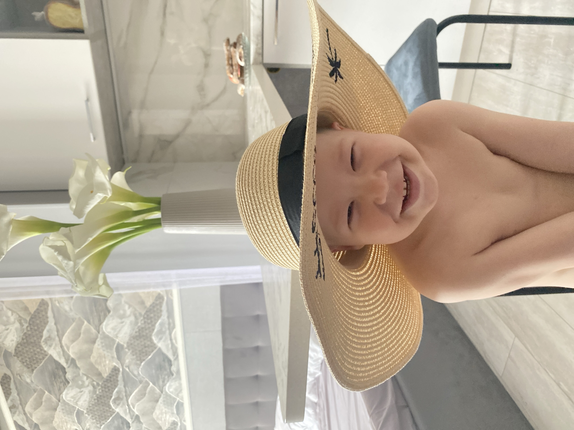 A young child, likely a toddler, is pictured wearing a large, straw sun hat that covers most of their face, though their smiling eyes and mouth are visible. The child is partially undressed, with their shoulders and chest bare. The hat is adorned with a black ribbon and a small embroidered insect design.
The child is in a brightly lit room, with a marble-patterned wall in the background and what appears to be a modern kitchen counter to the left. White calla lilies in a vase are also visible in the foreground. The setting suggests an indoor environment, possibly a home, with a relaxed and casual atmosphere.
The overall mood is cheerful and playful, emphasized by the child's wide smile. The lighting suggests it is daytime. The provided location context of Sochi, Russia, places the scene within a specific geographical setting, but no direct visual cues confirm this. There is no visible text in the image.