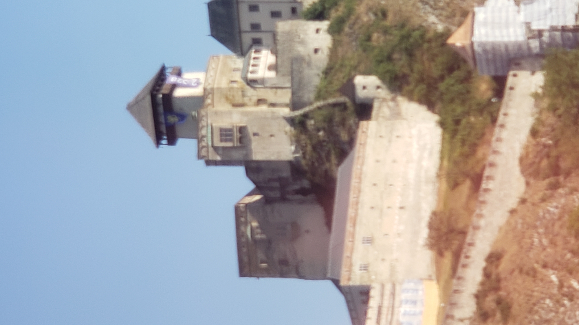 The image depicts Trenčín Castle, a historic fortress located on a prominent hill above the city of Trenčín, Slovakia. The castle complex, characterized by its stone architecture, includes several towers and fortifications. The main tower is topped with a dark, pyramidal roof and displays a banner with blue and gold elements. Adjacent buildings vary in height and architectural style, some featuring prominent windows and balconies. The castle is situated on a rocky, vegetated slope, with a lower wall and section of the city visible in the foreground. The sky is clear and blue.
