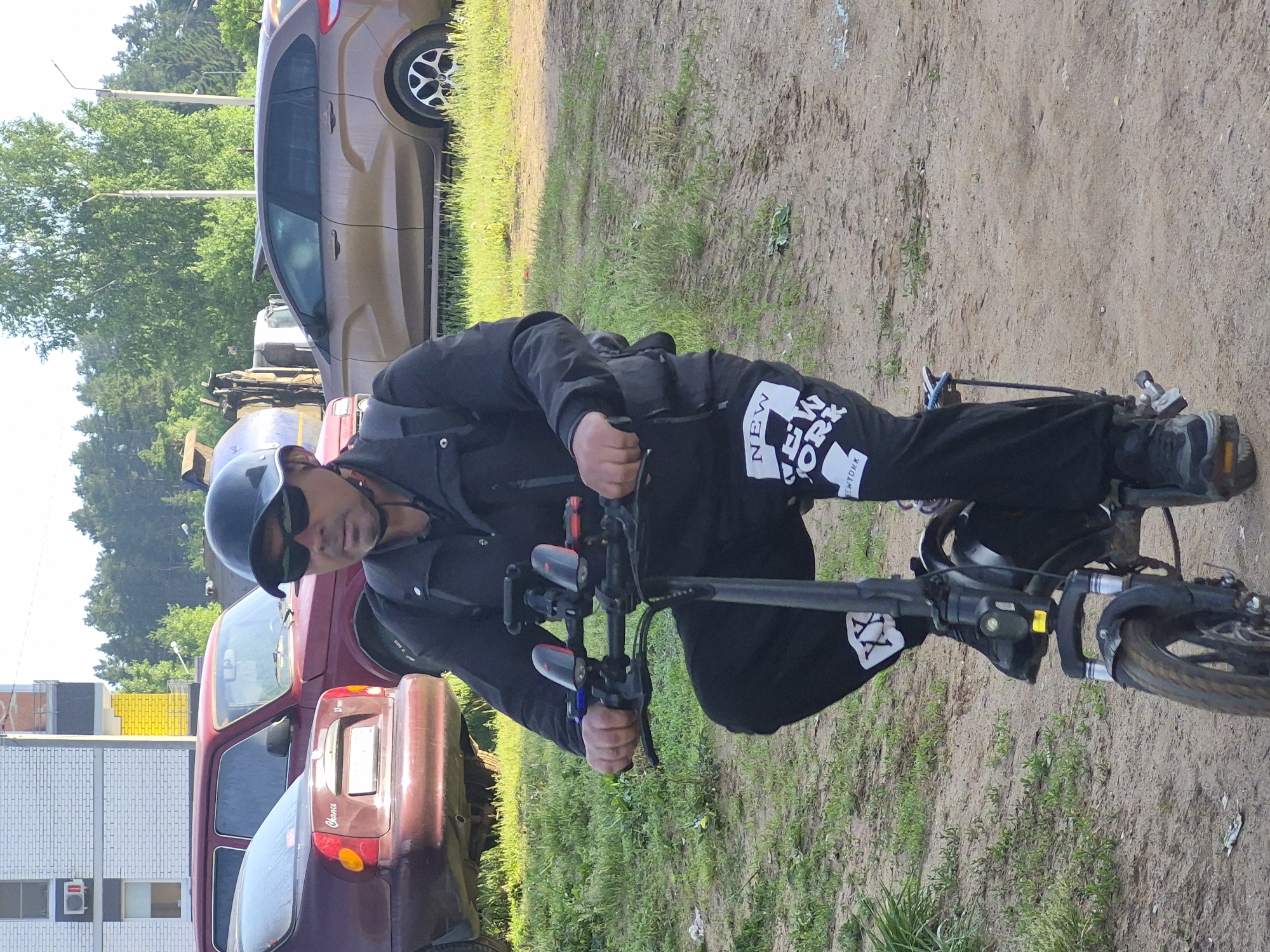 A man is depicted outdoors, straddling an electric scooter or bicycle on a dirt path. He wears a black helmet, dark sunglasses, a black jacket, and black pants. His left pant leg features white graphics, including the text "NEW YORK" and "NY" with two prominent "XX" symbols. He wears dark boots and grips the handlebars, looking directly at the camera with a neutral expression.
The setting is an unpaved, dusty area with patches of green grass. In the background, several vehicles are parked. A brown/gold sedan or small SUV is visible to the man's right, while a red car with "Chance" written on its trunk is behind him to the left. Other partially obscured vehicles and structures, including a building with white brick-like siding and a yellow wall, are further in the background. Abundant green trees line the distant horizon.
The scene is illuminated by bright daylight, suggesting clear weather, likely late morning or afternoon. The location appears to be a transitional area, possibly a rural road or the outskirts of a town, combining natural elements with man-made structures. The man appears to be paused during a ride.