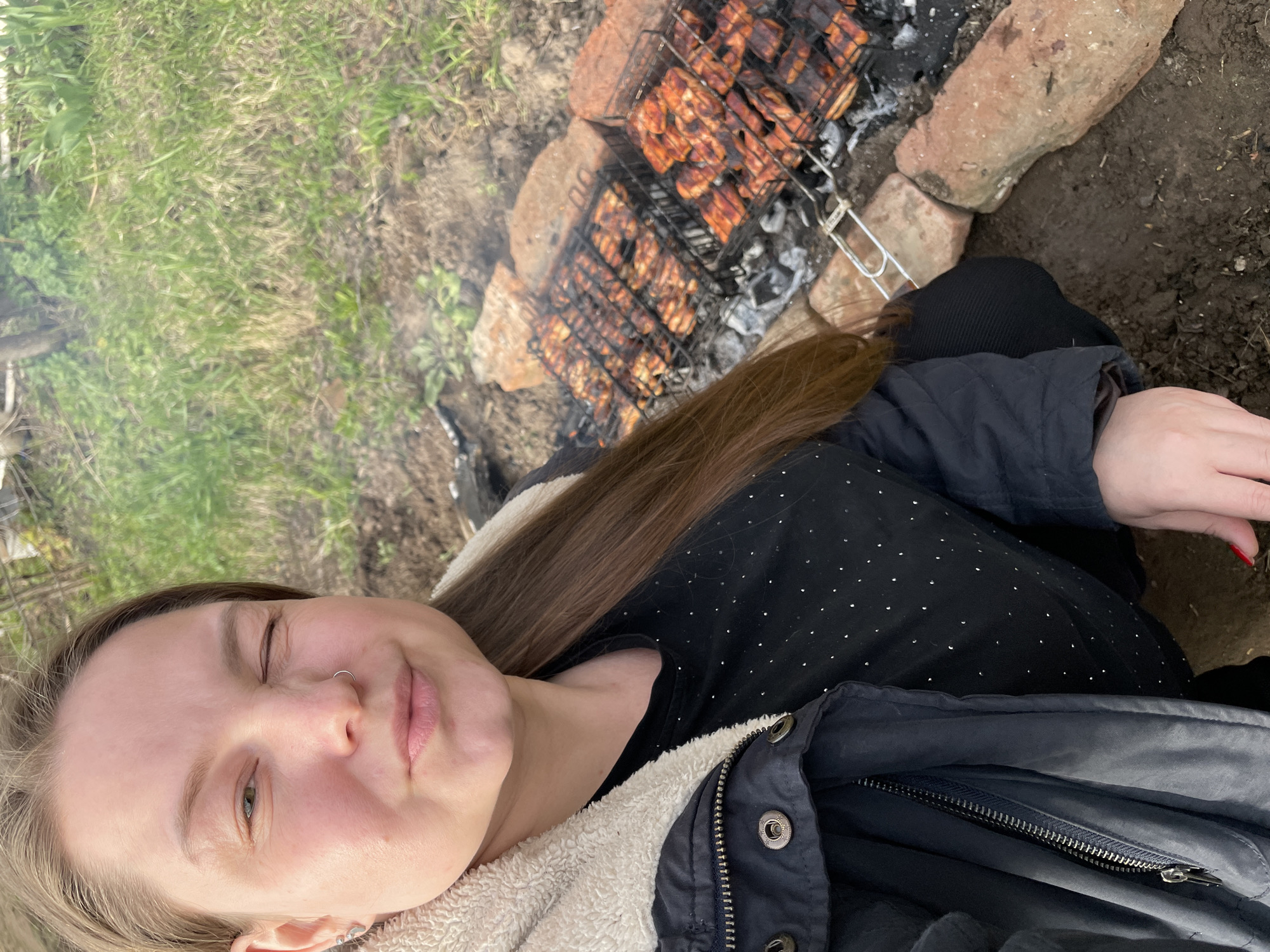 The image, rotated clockwise, captures a young woman taking a selfie while grilling outdoors in what appears to be a rustic setting.
The main subject is a light-skinned woman with long, straight brown hair, seen from the chest up. She is wearing a dark jacket with a cream-colored sherpa lining and a dark top adorned with small silver dots. She has a subtle smile and her eyes are partially closed, possibly due to smoke from the grill or bright daylight. A silver nose ring is visible, as are several small silver earrings on her left ear. Her left hand, with red nail polish, is also visible.
In the background, two grilling baskets filled with pieces of marinated meat, possibly chicken or pork, are cooking over hot coals. The grill setup is simple, with the coals contained by a few large stones on the ground. Wisps of smoke are rising from the cooking meat. The ground around the grilling area consists of patchy green grass and bare dirt.
The activity taking place is outdoor grilling. The scene suggests a casual daytime gathering or activity, likely in a rural or backyard environment. The overall atmosphere appears relaxed. There is no visible text in the image. Given the location context, this activity is taking place in an outdoor area in Балдейка, Russia.