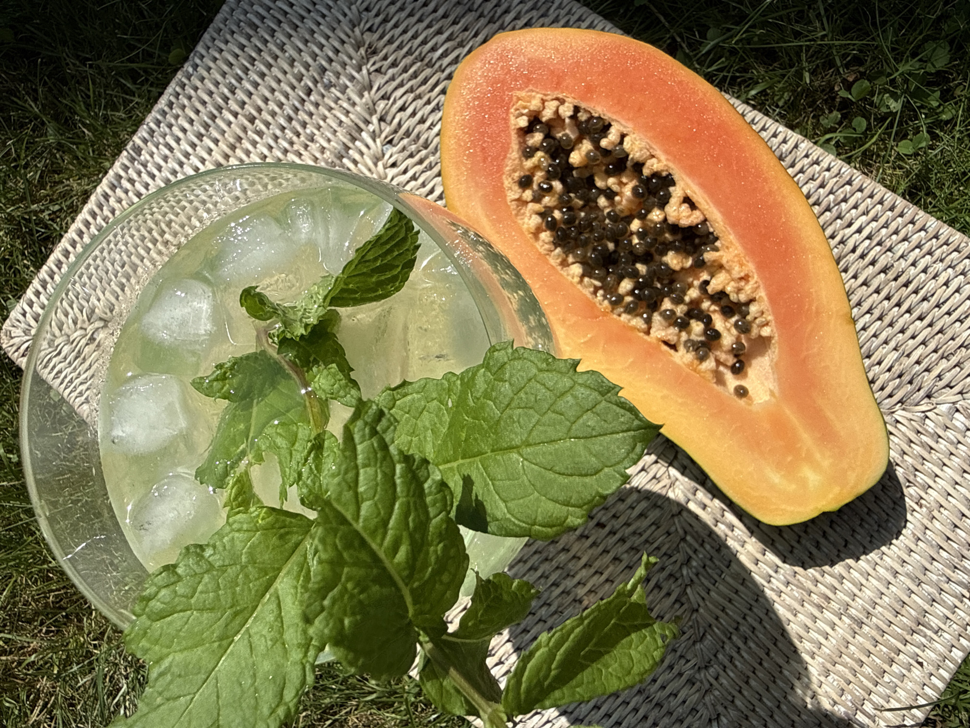 An overhead shot shows a refreshing beverage in a clear glass, filled with ice cubes and a sprig of mint. Beside it sits a halved papaya, its vibrant orange flesh and black seeds exposed. The arrangement rests on a woven placemat, which is placed on a patch of green grass. The scene is bathed in sunlight, casting shadows on the grass and placemat. The image does not depict any specific location in the city of Bad Sachsa, Germany.