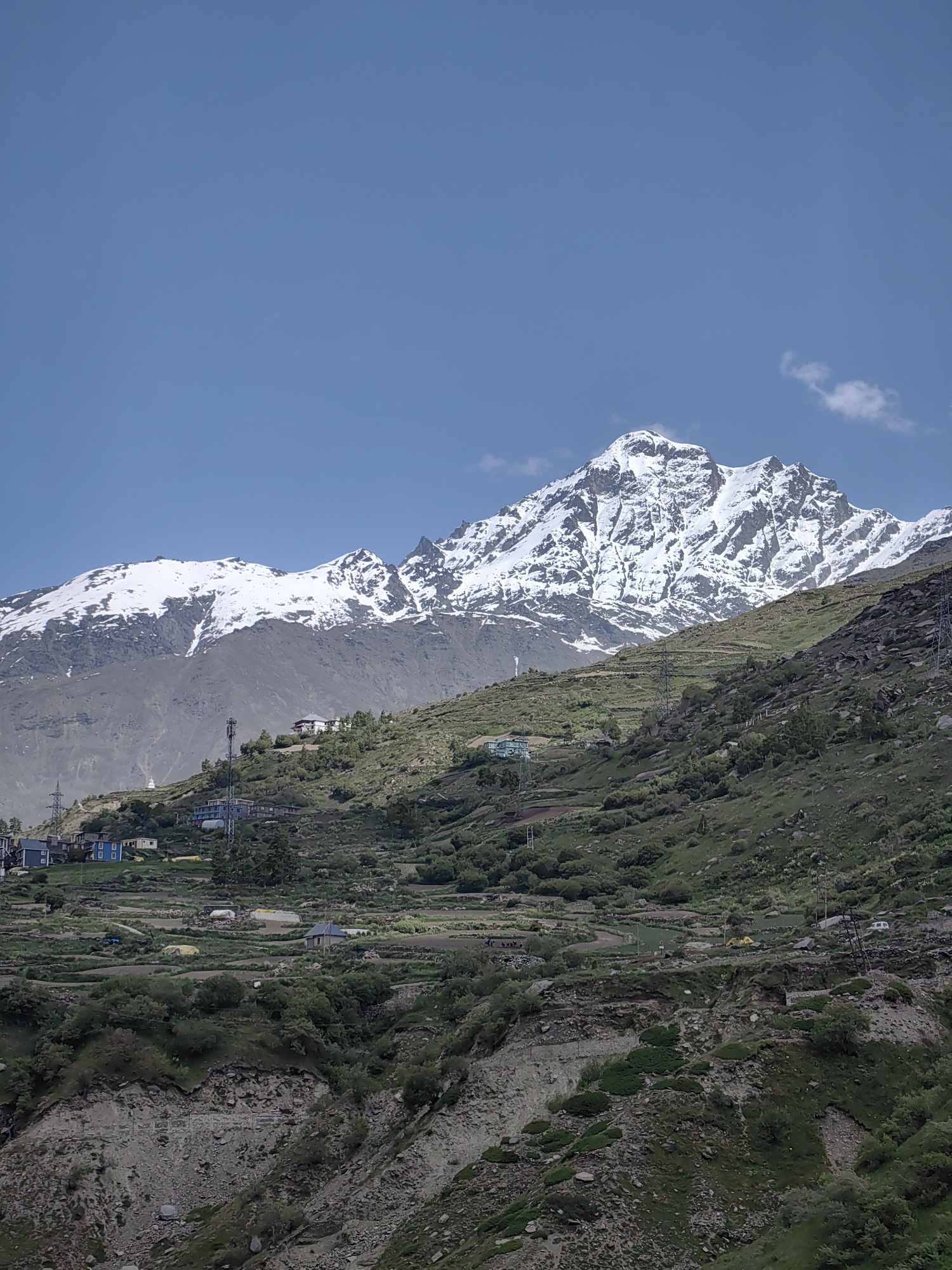 The image depicts a vast, snow-capped mountain range under a clear blue sky. The lower slopes of the mountains are covered in green vegetation, with terraced fields and scattered buildings forming a small settlement. Power lines and transmission towers are visible on the slopes, indicating the presence of infrastructure. The terrain is rugged and rocky, with evidence of erosion. The overall scene conveys a sense of natural beauty and human habitation in a mountainous region. The location is identified as a village in the Indian Himalayas.