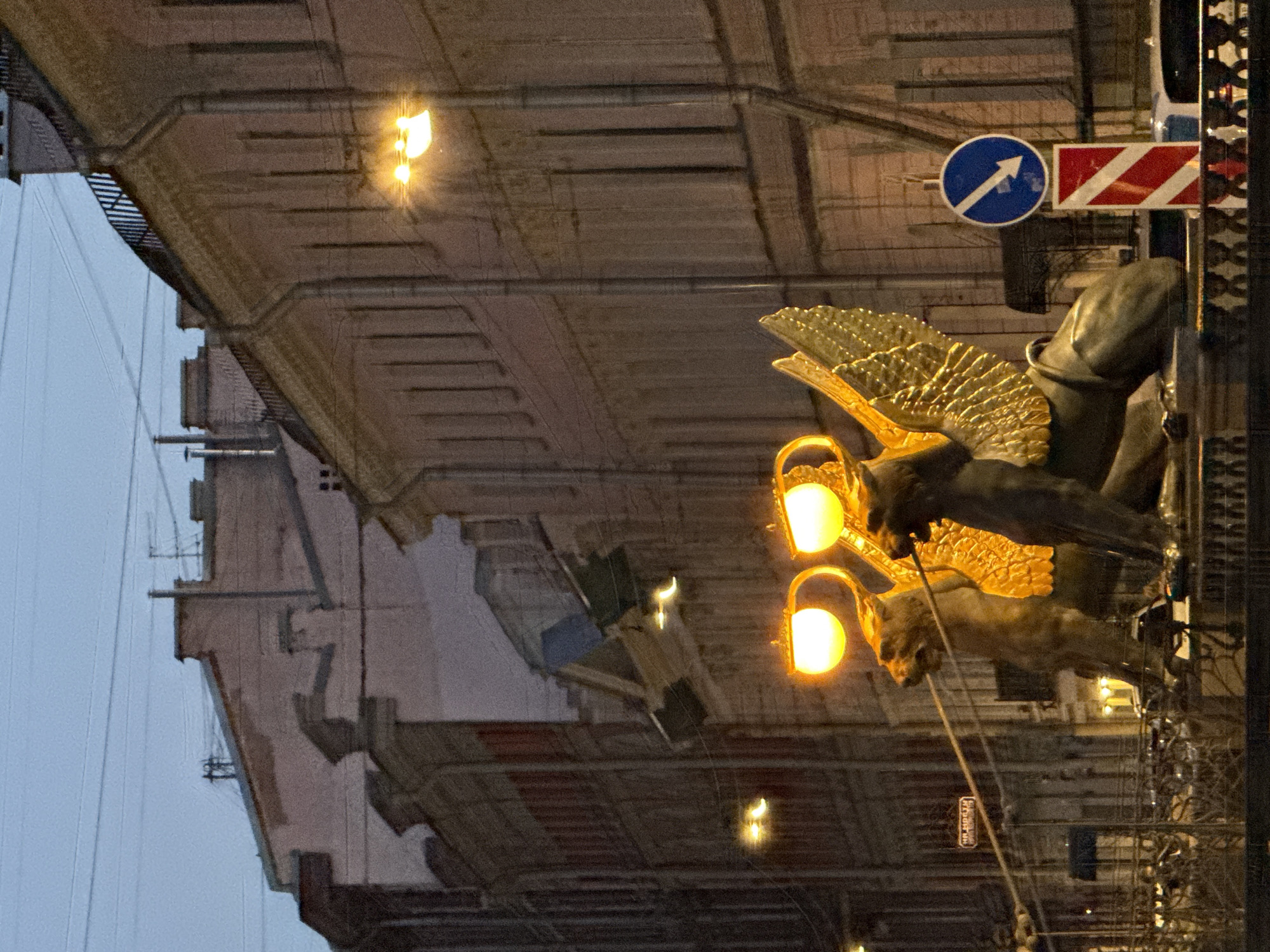 The image depicts a night scene in Saint Petersburg, Russia, featuring the griffins of the Bank Bridge. Two bronze griffins with large, gilded wings stand on pedestals, their bodies facing forward and heads turned slightly. Each griffin is adorned with two illuminated lamps, casting a warm glow. The griffins are positioned on the Bank Bridge, which is visible in the foreground with ornate metal railings. Behind the griffins, the architecture of Saint Petersburg lines the street, with multi-story buildings featuring classical facades and illuminated windows. A blue circular traffic sign with a white arrow pointing down and to the left is visible to the right of the griffins, along with a red and white striped directional marker below it. The sky is dark, indicating it is evening or night. The overall impression is a historic and atmospheric urban landscape.