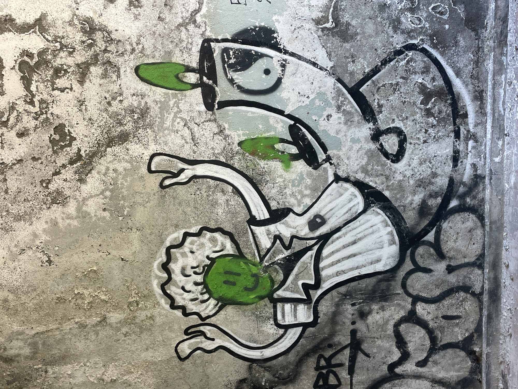 A colorful graffiti artwork adorns a weathered concrete wall in Abaata, Abkhazia. The central figures are a cartoonish character with a green, smiling face, wearing a white chef's hat and a striped shirt. To its right is what appears to be a spray paint can, also with a green element on top, and a yin-yang symbol on its side. The artwork is rendered in bold black outlines with splashes of white and green paint. The wall itself is textured and shows signs of age and decay, with patches of peeling paint and grime. Below the main artwork, more graffiti is visible, including black spray-painted lettering. The overall scene depicts an urban art installation in a state of disrepair, typical of many public spaces.