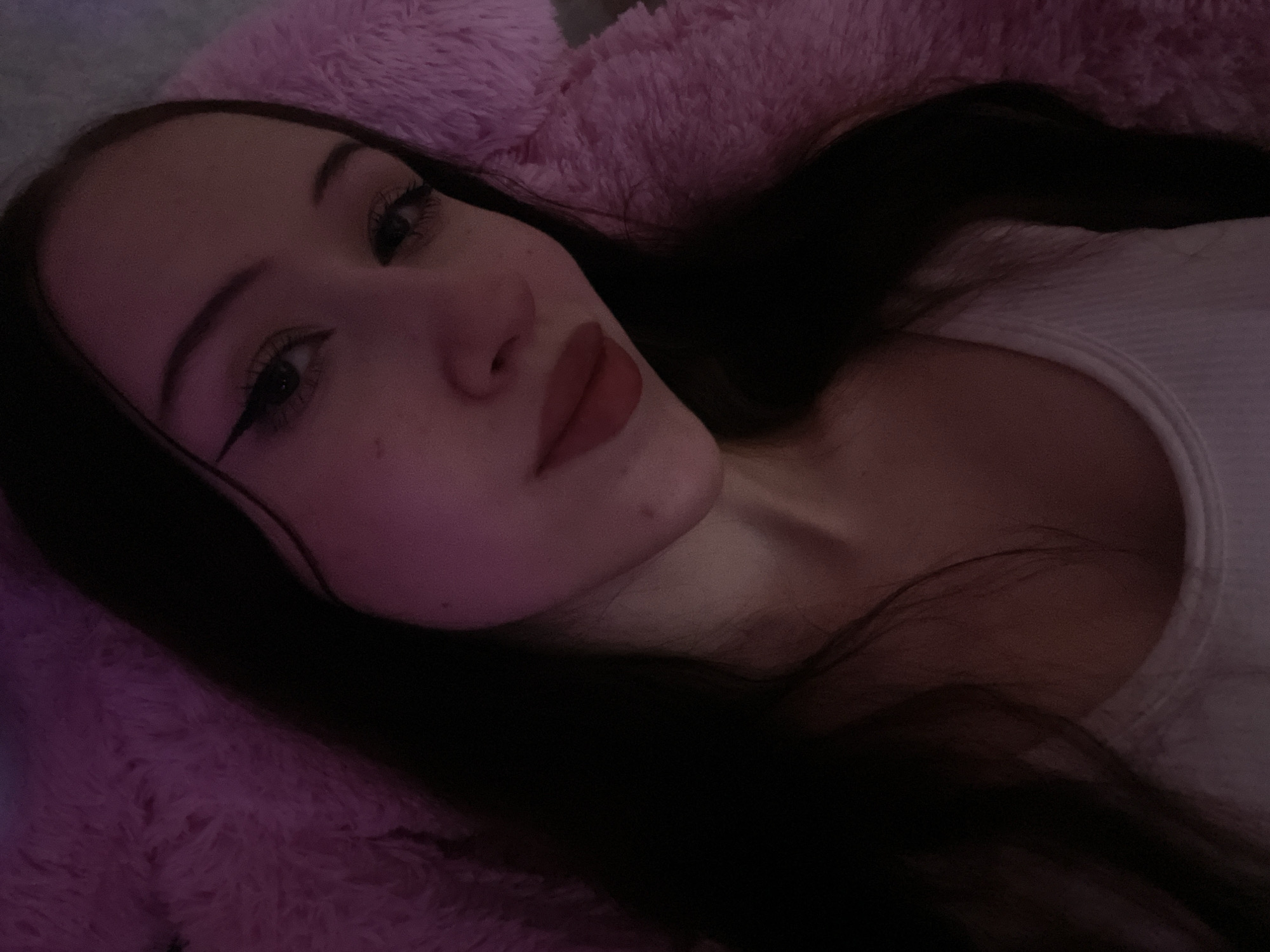 This is a close-up, low-angle selfie of a young woman with long, dark hair, wearing a white tank top. She is lying down on a fluffy pink surface, possibly a bed or a blanket. The lighting is dim and pink-toned. Her makeup includes winged eyeliner and red lipstick. There is a mole visible on her cheek. The image is taken indoors, and no specific location in Votkinsk, Russia can be determined from the visual information.