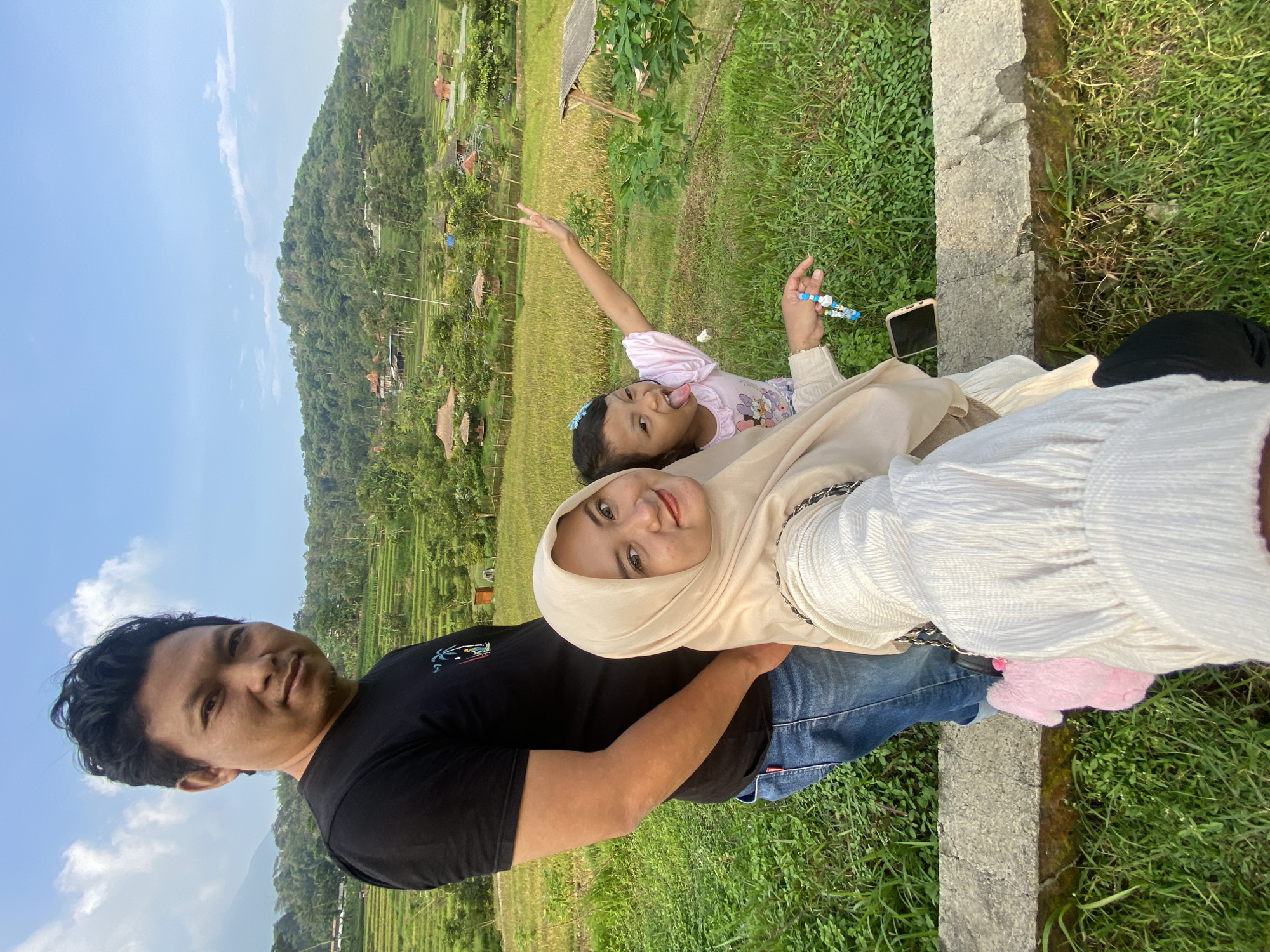 A daytime, eye-level, color photograph shows three individuals near Penanggungan, Indonesia. A man with black hair and a black t-shirt is on the left. A woman wearing a cream hijab and white long-sleeved shirt is next to him. A young girl in a pink shirt is reclining between them. The woman is smiling and looking at the camera. The girl has her arm up, gesturing. A smartphone lies on the concrete wall behind her. In the background are rice paddies and traditional Indonesian houses. A forested mountain is visible in the distance under a blue sky.