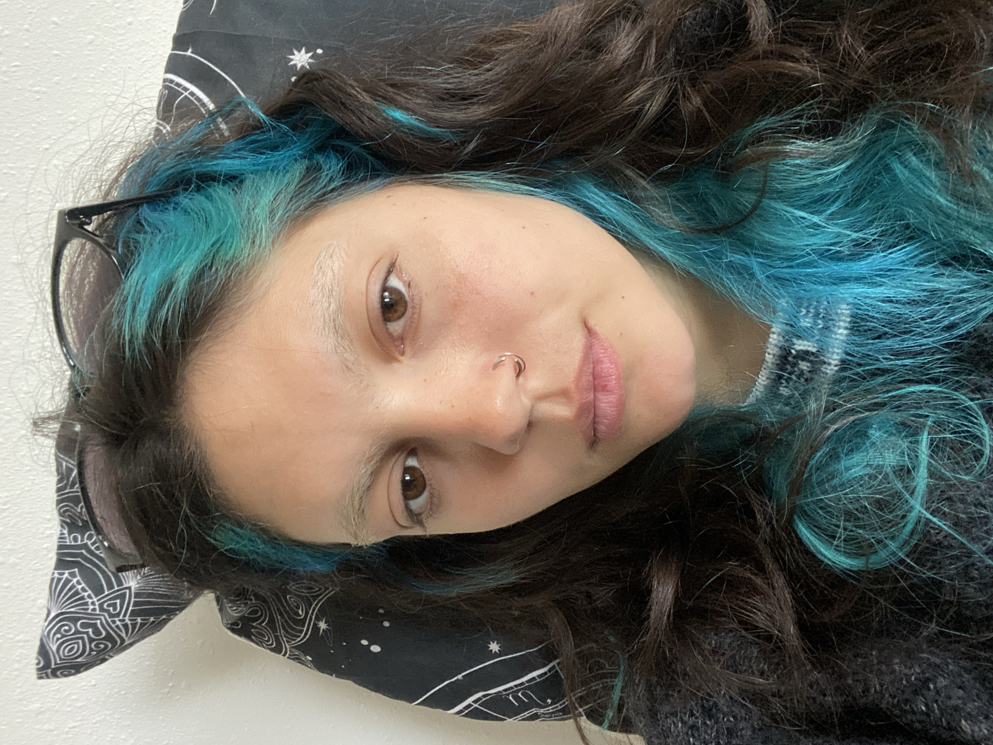 A close-up, tilted, selfie-style image of a person with brown hair and dyed blue streaks. The person has fair skin, brown eyes, and a nose ring. Black-framed glasses are visible near their face, resting against a pillow. The pillowcase is black with white celestial designs. The person is wearing a dark gray sweater or top. The background consists of a white wall.
The location in the city of Gijón/Xixón, Spain is unknown.