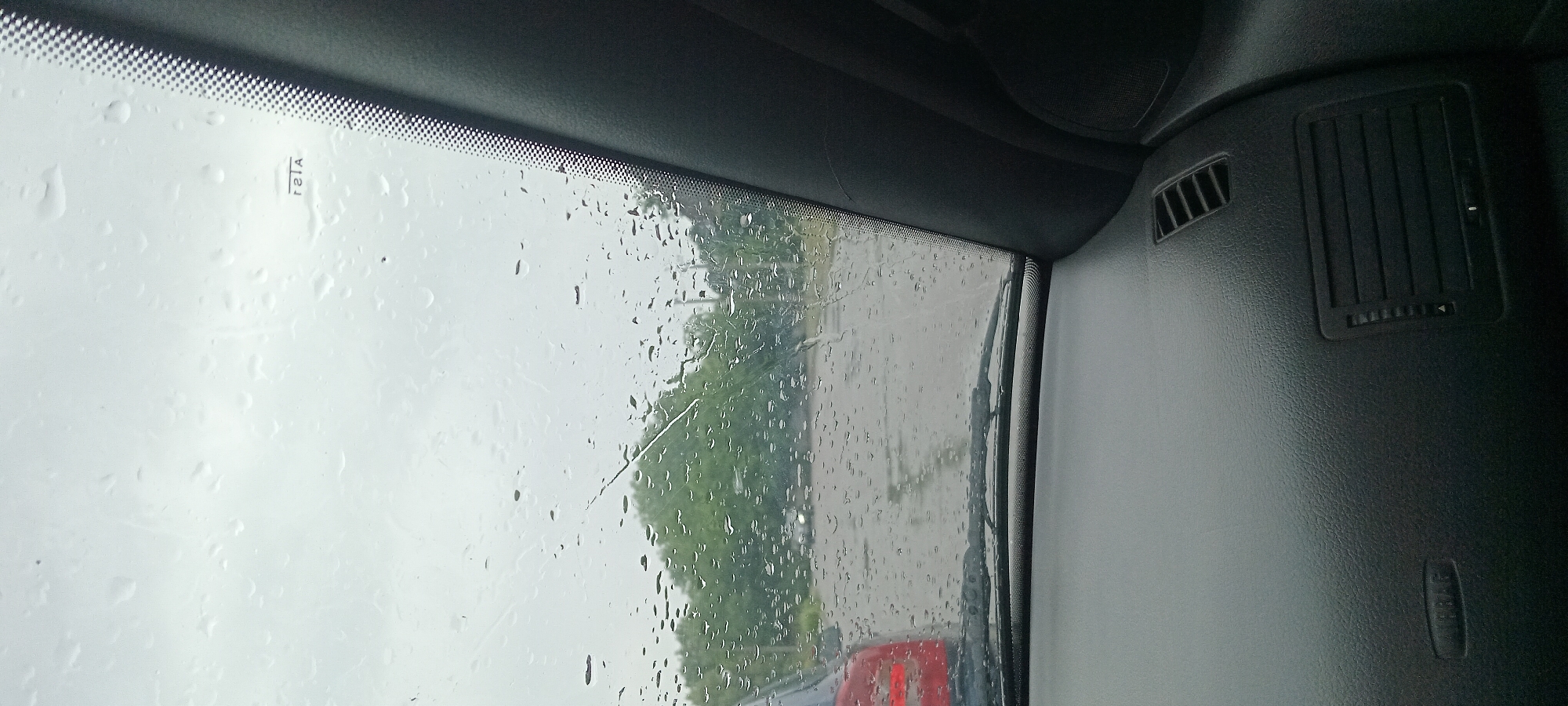 The media file captures a view from inside a vehicle, looking out through a rain-streaked windshield. The weather is clearly overcast and rainy, with numerous water droplets distorting the view of the outside scene. The exterior shows a blurry landscape of trees and what appears to be a road with another vehicle in front, indicated by a red taillight. The scene is likely taking place during the daytime due to the ambient light, but the overcast conditions suggest a muted light.
Inside the vehicle, the dashboard is visible, featuring a black textured surface. There are two air vents present; one smaller one on the left and a larger one on the right. A subtle detail is the text "AIRBAG" embossed on the dashboard, suggesting the vehicle's interior. There is also a small rectangular sticker on the windshield with vertical black lines and the text "r2tA" in black. The overall mood conveyed is one of being on a journey during inclement weather, with the focus on the obstructed view and the interior of the car. The location context of Koviahy, Ukraine, doesn't provide specific visual cues within the image itself.