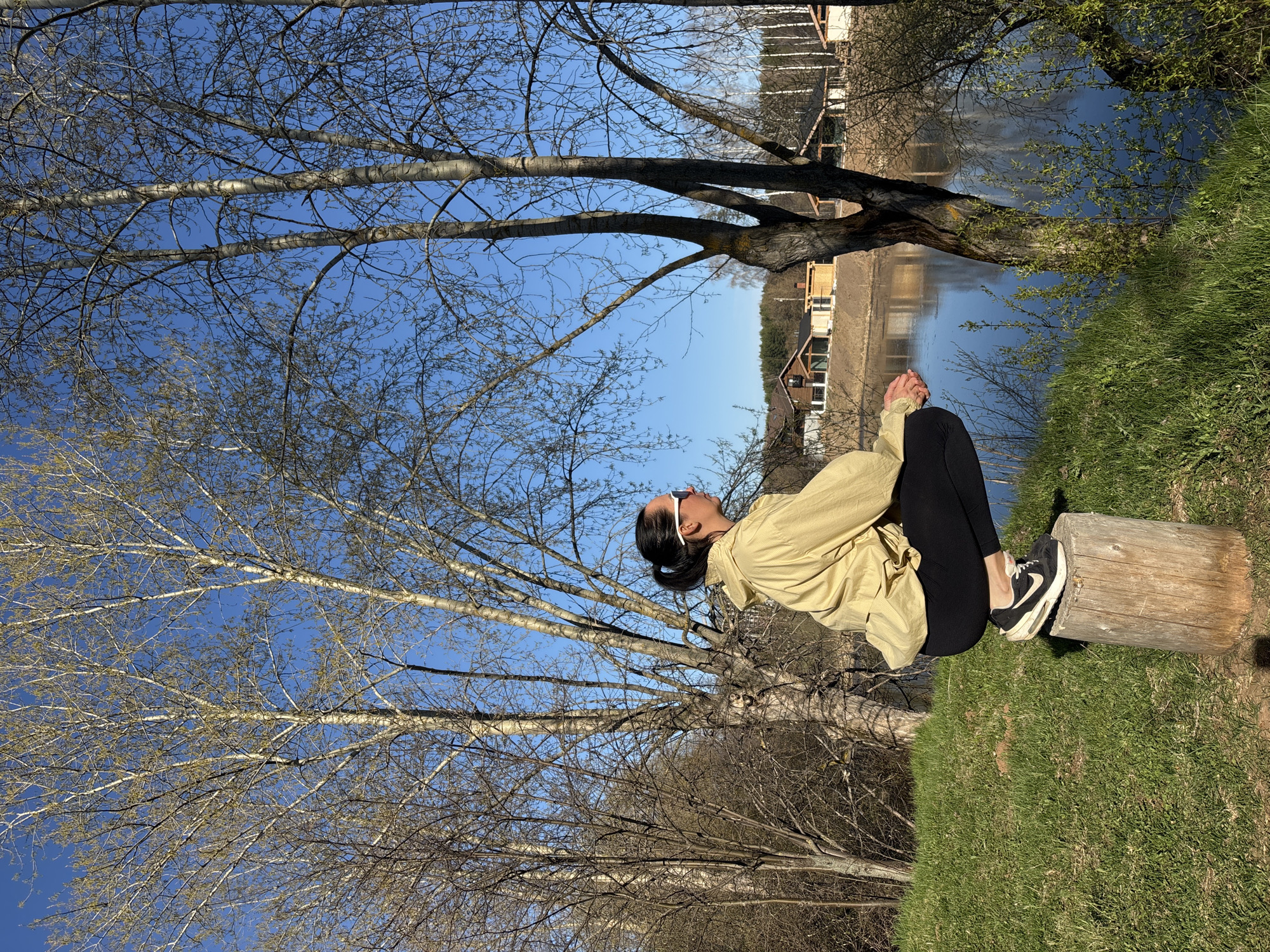 A woman is seated on a log on a grassy area. She is wearing black pants, black and white shoes, a light-colored jacket, and white sunglasses. The backdrop features a body of water with buildings on the opposite shore, and trees. The setting is outdoors. The photo was taken in the city of Chaikovsky, Russia.