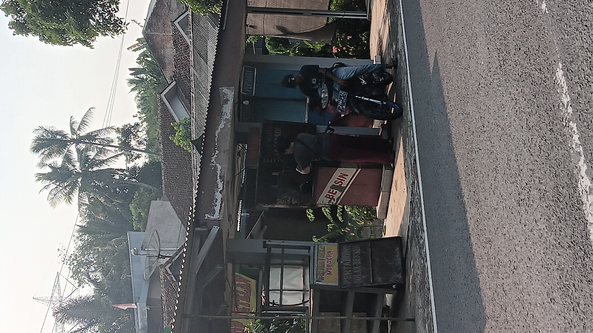 The image depicts an outdoor scene, likely in Tanjungsari, Indonesia, featuring a small roadside establishment that appears to be a gas station or convenience store. In the foreground, a paved road with a white dividing line runs across the bottom of the frame. To the right, a motorcycle is parked, with a man wearing a black t-shirt and light-colored pants seated on it, facing the establishment. To the left of the motorcycle, a person in a dark t-shirt and a long, maroon garment is interacting with a fuel dispenser. The fuel dispenser is red and white, with the word "BENSIN" (Indonesian for gasoline) visible. Behind the fuel dispenser, there is a structure with a corrugated metal roof, possibly a covered awning or part of the building. A sign with yellow text that reads "TOP UTELAN" is visible on the left side, above a wooden counter or shelf. Another sign, with yellow text that says "MERDEKA" and below it "PERTAMINA SOLAR BENSIN," is placed on a dark board. The background shows trees, some of which are palm trees, and houses with tiled roofs. A tall transmission tower is visible in the far left background. The sky is overcast or hazy.
