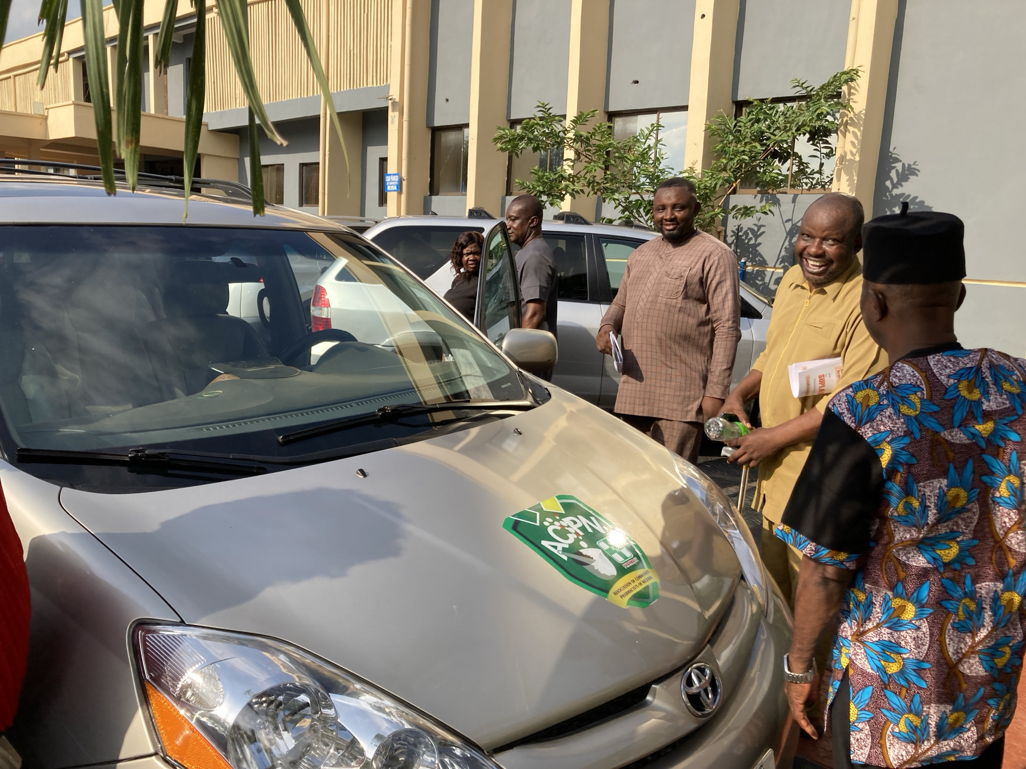 A group of men and one woman are gathered outdoors in what appears to be a parking area in Umuahia, Nigeria. In the foreground, a light-colored Toyota Sienna minivan is parked, facing towards the right. A prominent green and yellow sticker on the hood of the minivan displays the acronym "ACPN" and the text "ASSOCIATION OF COMMUNITY PHARMACISTS OF NIGERIA."
Several individuals are visible around the vehicle. On the right, a man wearing a black cap and a colorful, patterned shirt with blue, yellow, and brown floral designs is seen from behind, facing another man. This second man, wearing a light brown or tan long-sleeved shirt, is smiling broadly and holding a plastic bottle and some papers with partially visible text "SUPPL...". He appears to be engaging in conversation. To his left, another man in a plaid brown shirt is also smiling, holding papers, and looking towards the right. Further in the background, near a silver car, a man in a dark grey t-shirt stands near an open car door, and a woman in a black top is partially visible inside the car.
The background features a modern-looking building with beige and grey facades and rectangular windows. Green foliage and small trees are visible between the building and the parked cars. The scene is brightly lit by natural sunlight, casting visible shadows, indicating it is likely midday or early afternoon. Palm fronds hang into the frame from the top left, suggesting a warm climate. A partially obscured blue sign in the background reads "NO PARKING...". The overall mood appears friendly and social.