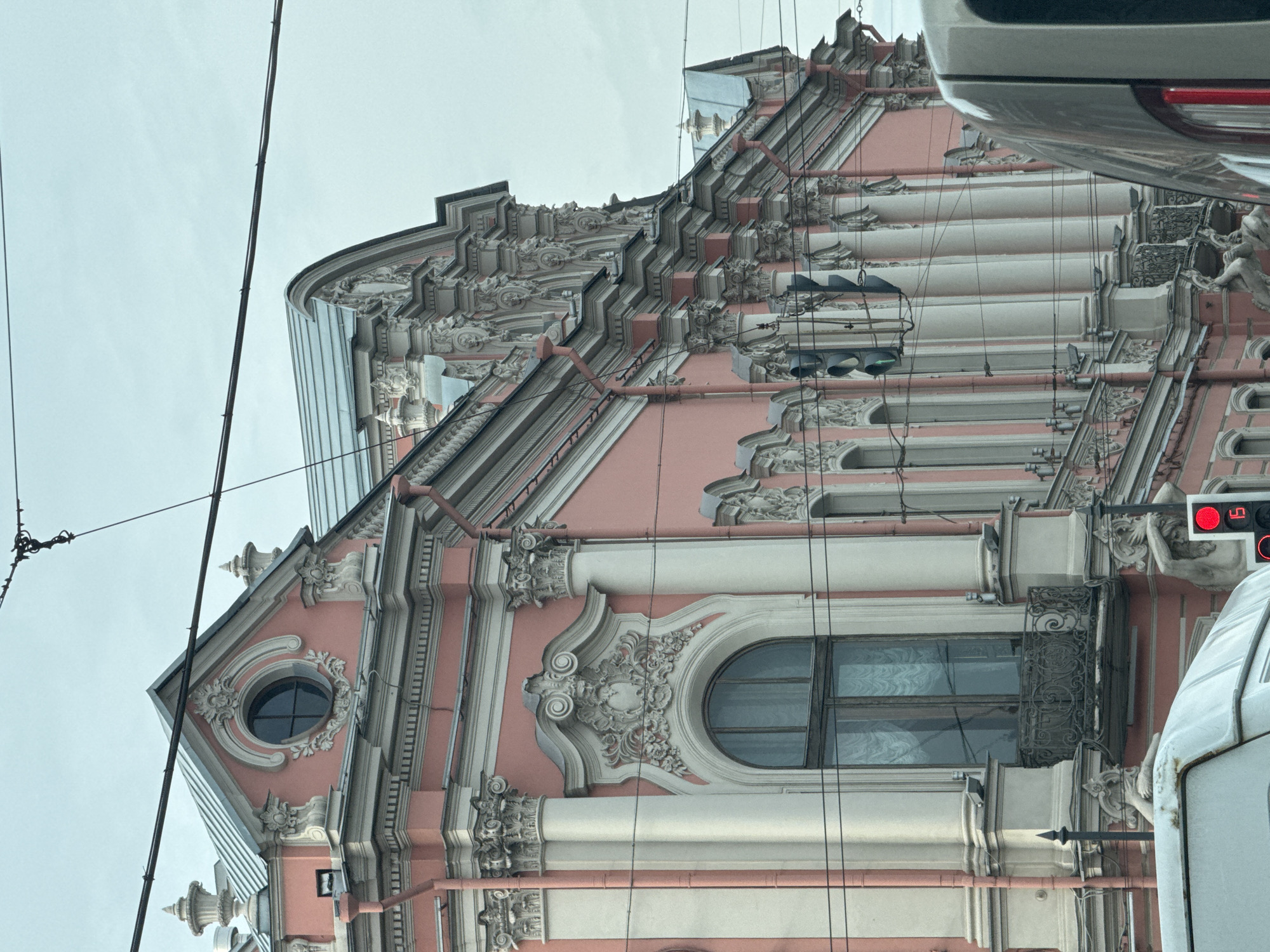 The image shows a detailed view of the architecture of the Eliseyev Emporium, located at 56 Nevsky Prospect in Saint Petersburg, Russia. The building features a pink and white facade adorned with ornate details, including sculptural elements, columns, and decorative moldings. Arched windows and a round window on the corner emphasize the building's Beaux-Arts style. Electrical wires run across the front of the building, and a traffic light is visible on the right, indicating that the photo was taken from street level. Part of a vehicle is visible on the right side of the image. The sky is overcast.