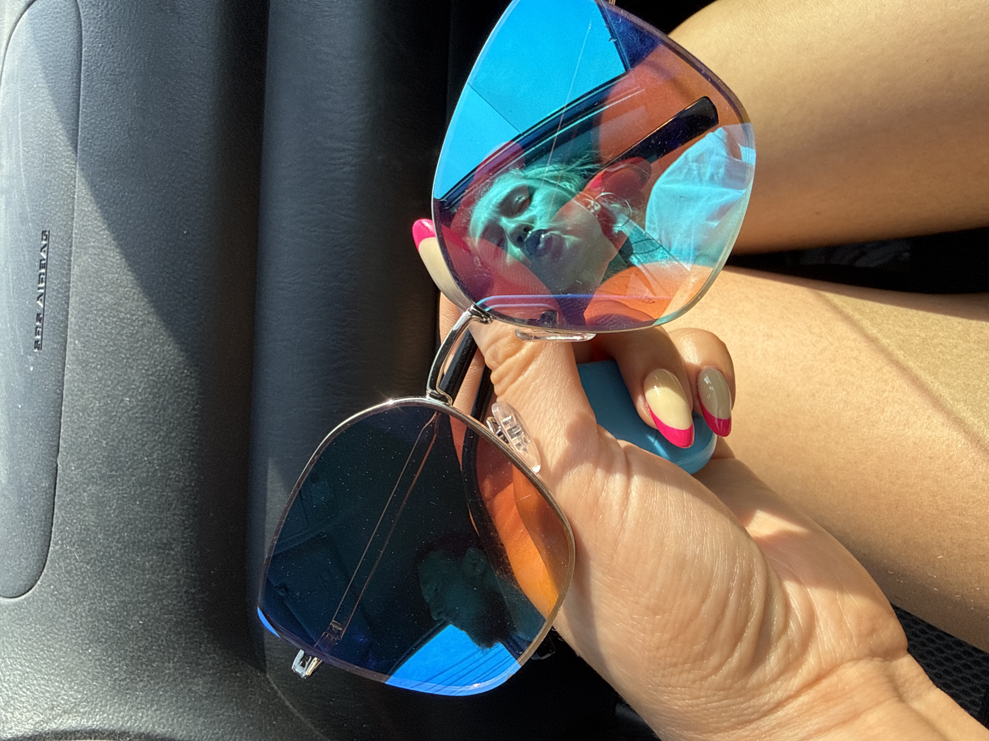 A hand is holding up a pair of sunglasses, with a reflection of a person's face in the lenses. The hand has manicured nails with a nude color and pink tips. The sunglasses have a blue tint and a silver frame. A car's dashboard is visible on the left side of the image, and a leg is visible on the right. The location is not identifiable as Chaikovsky, Russia, from the information in the image.