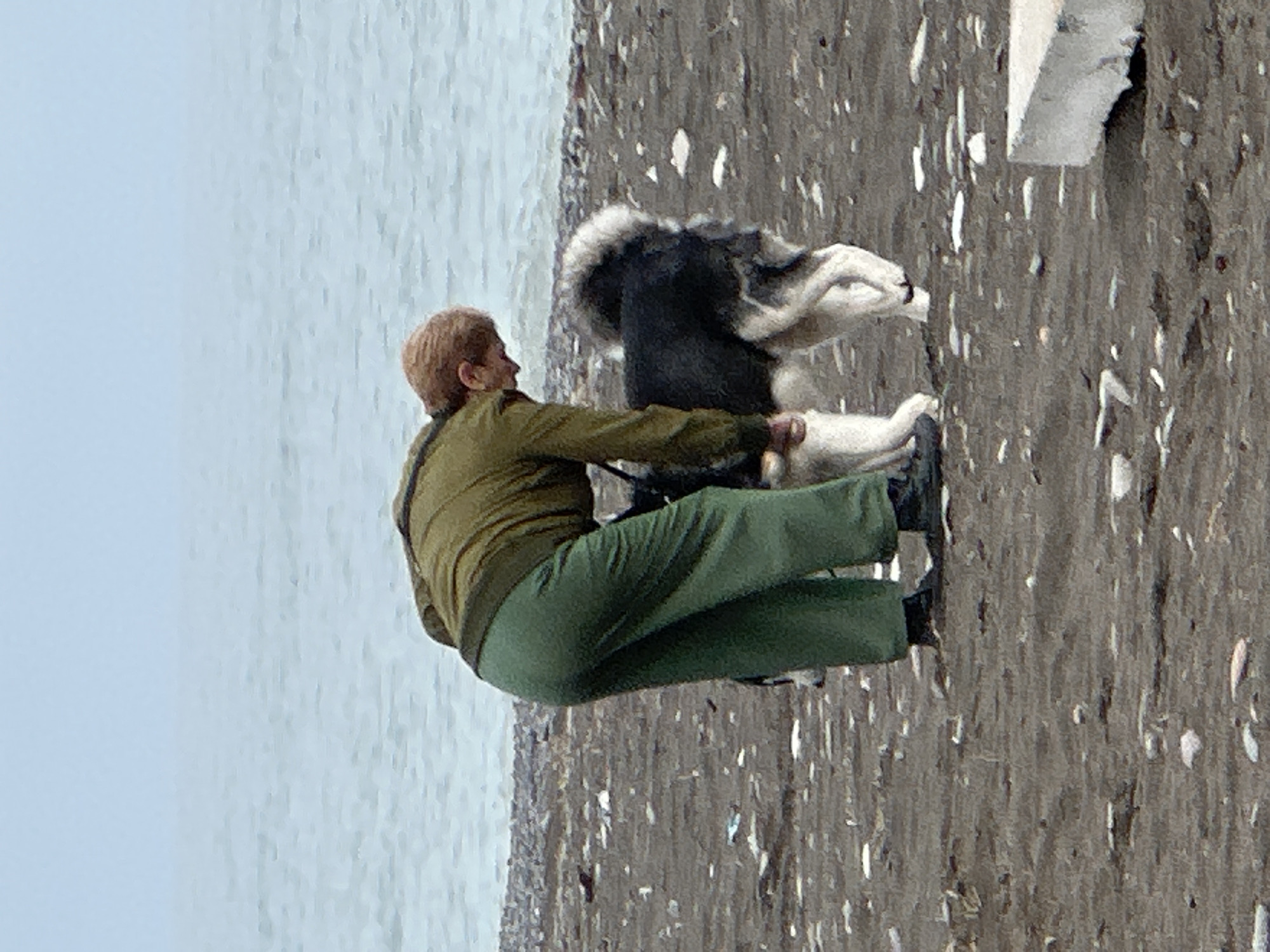 An outdoor scene shows a person sitting on a dark, pebbly surface near a body of water, interacting with a black and white dog. The person is wearing an olive green jacket and matching pants with black shoes. The dog is positioned near the person's legs and appears to be engaging with them. The setting suggests a beach or shoreline location, with the sea visible in the background and a rocky or pebble-covered ground. The location of the image cannot be determined within the city of Batumi, Georgia.