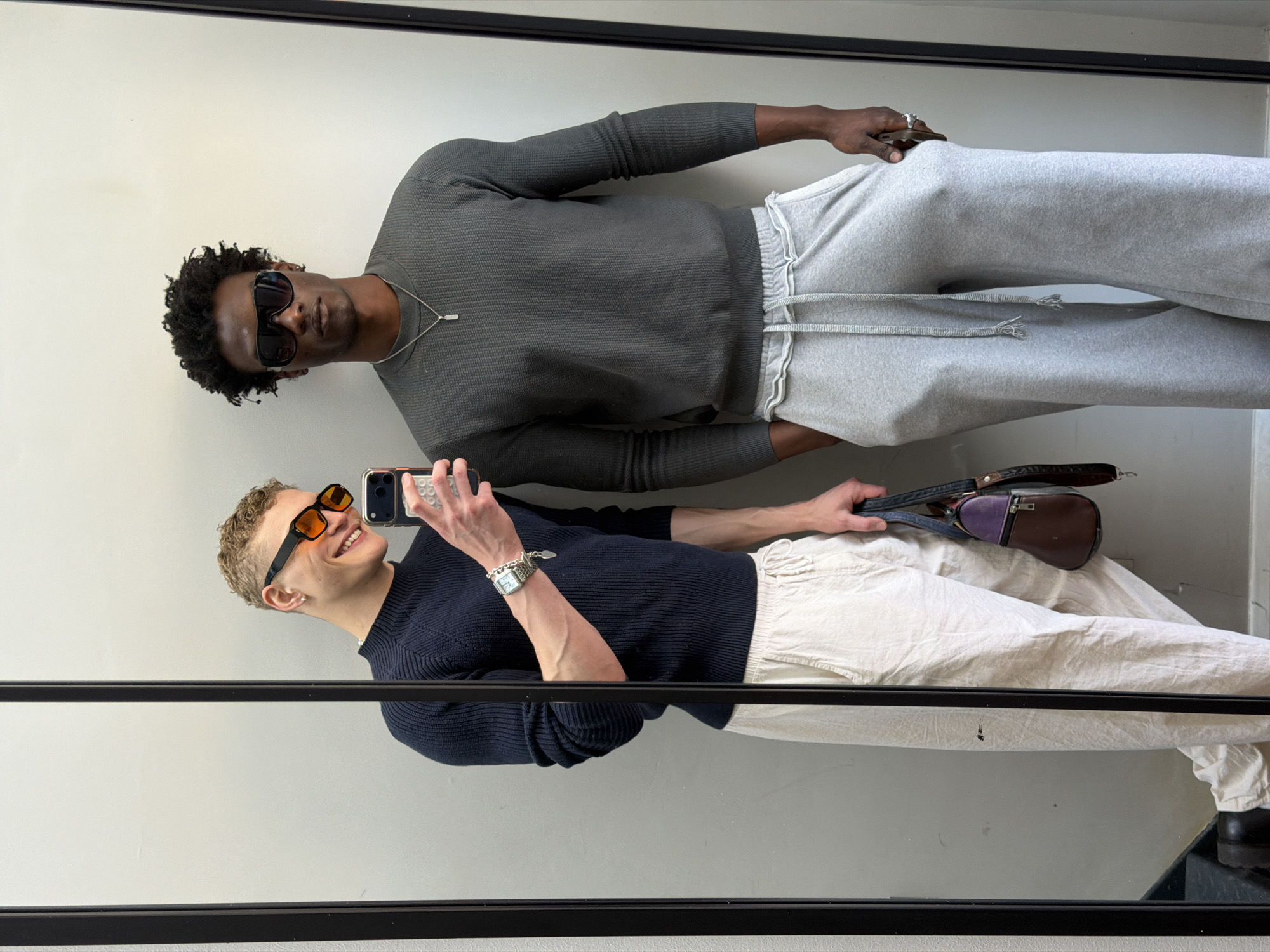 Two people are posing in front of a mirror. The person at the top wears a grey long-sleeved shirt, grey sweatpants, sunglasses, and a silver necklace. They have dark skin and curly hair. The person at the bottom wears a dark blue long-sleeved shirt, cream-colored pants, orange sunglasses, and a silver watch. They are holding a phone and a small handbag. The background consists of a white wall and a black frame around the mirror. The setting appears to be indoors, possibly a dressing room or retail space, and the location cannot be determined as London, United Kingdom, from the image.