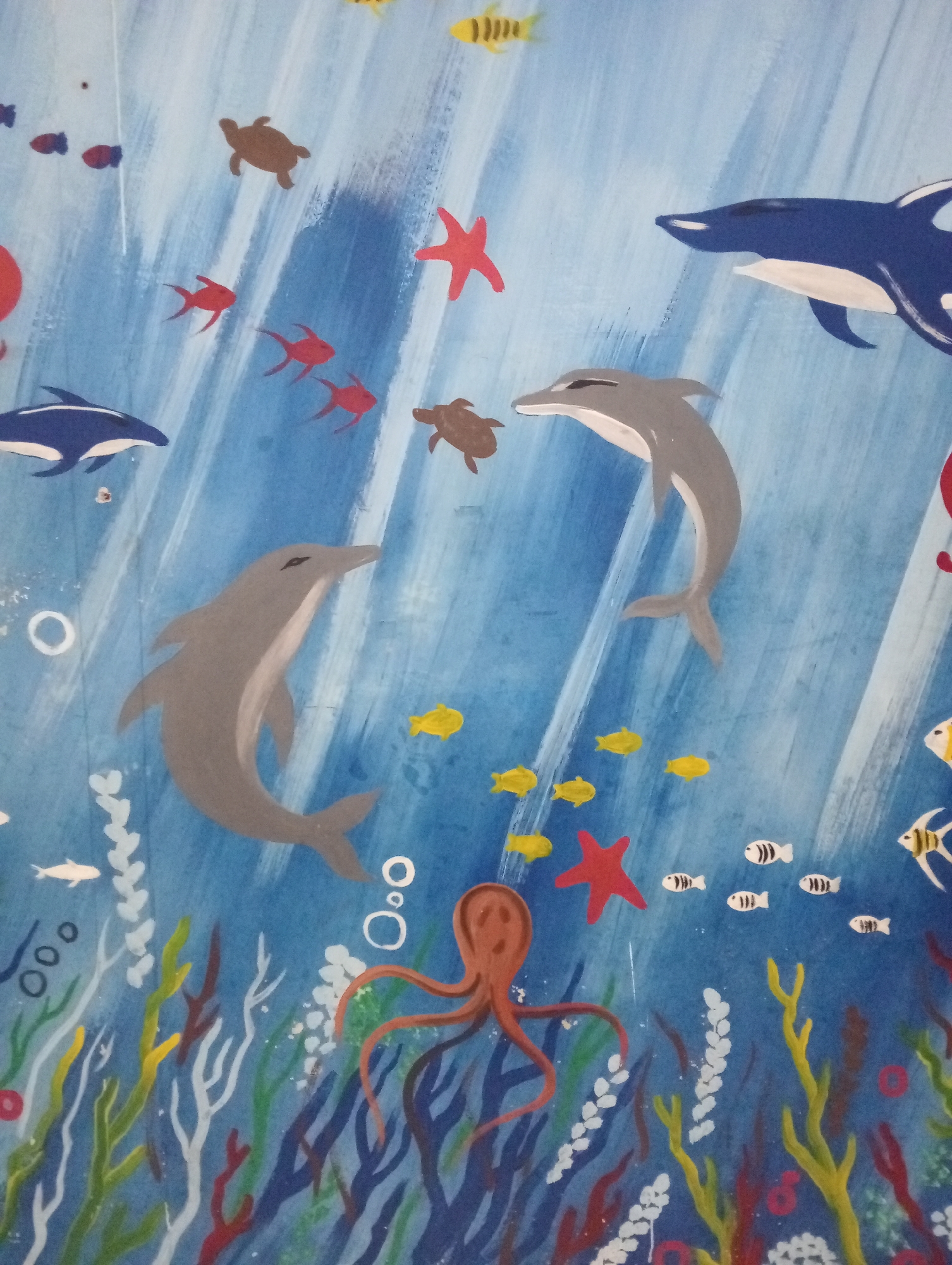 The image is a painting of an underwater scene, likely a mural on a wall. It depicts various marine life such as dolphins, fish, turtles, starfish, and an octopus swimming amidst colorful coral and seaweed. The water is depicted with brushstrokes suggesting currents. There is no specific indication of this scene being located in Pringsewu, Indonesia, beyond what has been provided as context.