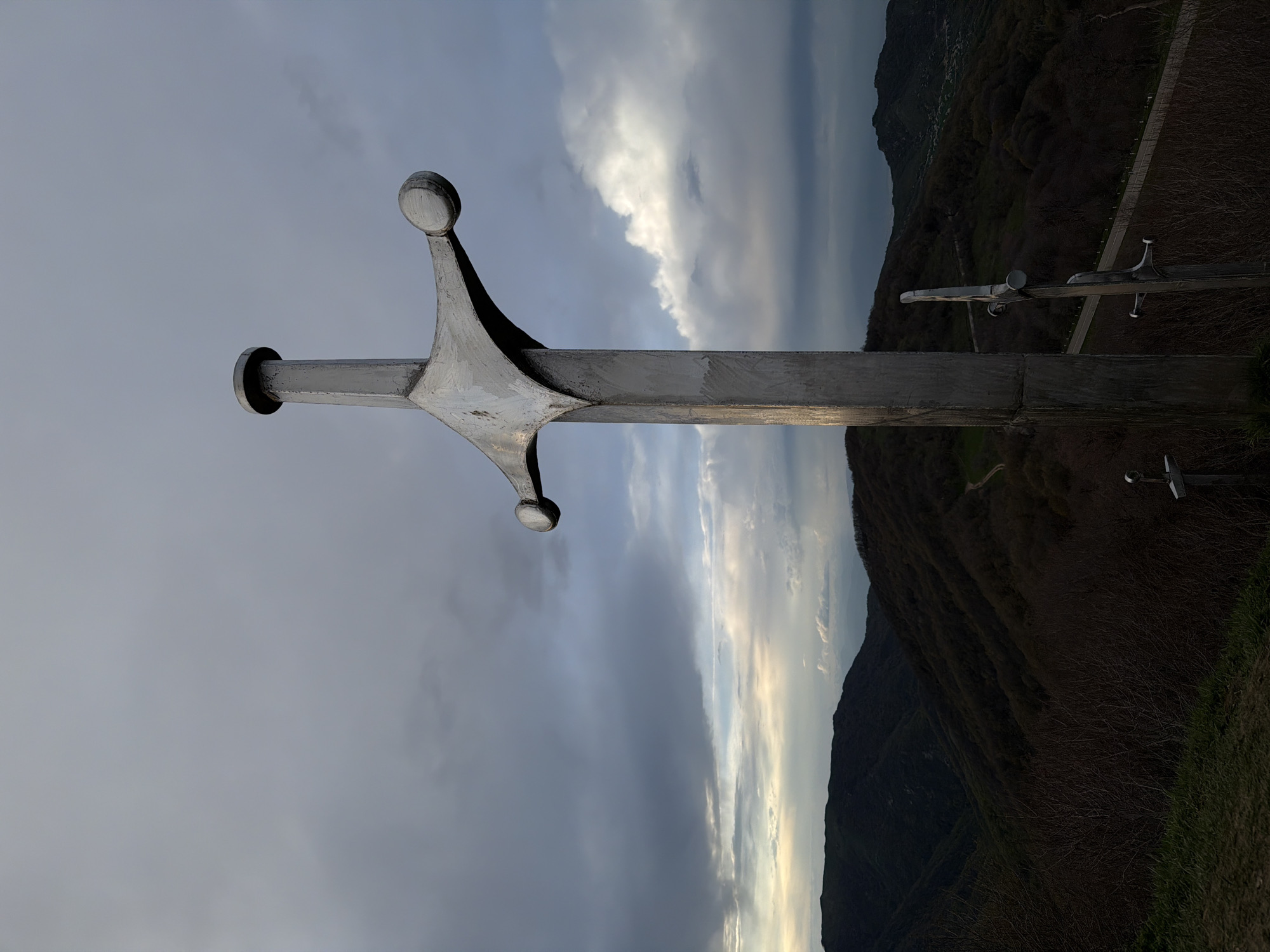 A low-angle, outdoor photograph depicts a large, metal sword monument set against a backdrop of a cloudy sky and a mountainous landscape in Zemo Nichbisi, Georgia. The sword's hilt features rounded pommels and a prominent cross-guard. Further down a metal path are two smaller metal swords. The environment consists of dark, tree-covered mountains under a sky with visible clouds and varying light conditions.