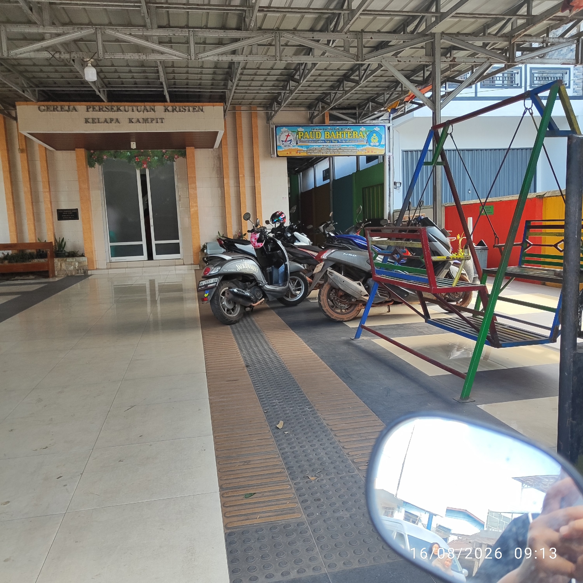 The image depicts the entrance to a building with a sign overhead that reads "GEREJA PERSEKUTUAN KRISTEN KELAPA KAMPIT," indicating it is the Christian Fellowship Church of Kelapa Kampit. Adjacent to the church entrance, a blue sign advertises "PAUD BAHWERA," suggesting a kindergarten or early childhood education center. Several motorcycles are parked in the covered area in front of the building. To the right, a colorful metal swing set is visible, partially obscuring some of the motorcycles. The ground is tiled, with a section of textured paving tiles running along the pathway. In the foreground, a motorcycle's rearview mirror reflects the sky and some buildings, along with a date stamp reading "16/08/2026 09:13". The overall scene suggests a public space, likely in an urban or semi-urban setting in Indonesia, specifically the city of Pembaharuan, based on the church's name.