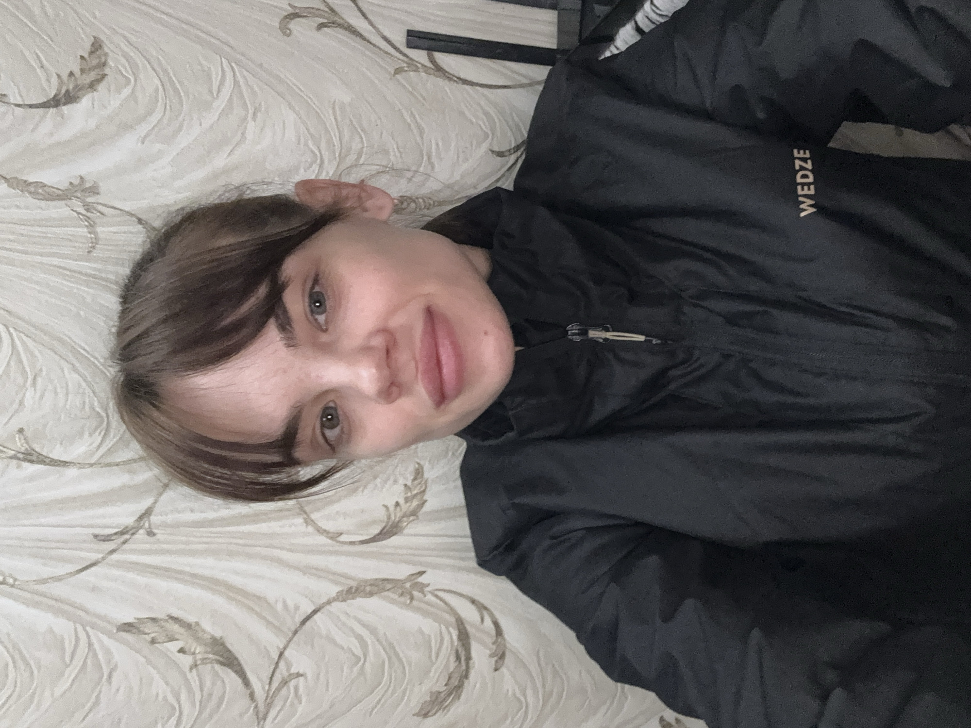 A woman with short brown hair and fair skin is lying on a patterned surface, possibly a bed or couch, in a selfie-style photograph. She is wearing a black jacket with the brand name "WEDZE" visible on the right side. The background features a floral patterned texture. Location is not apparent from the image.
