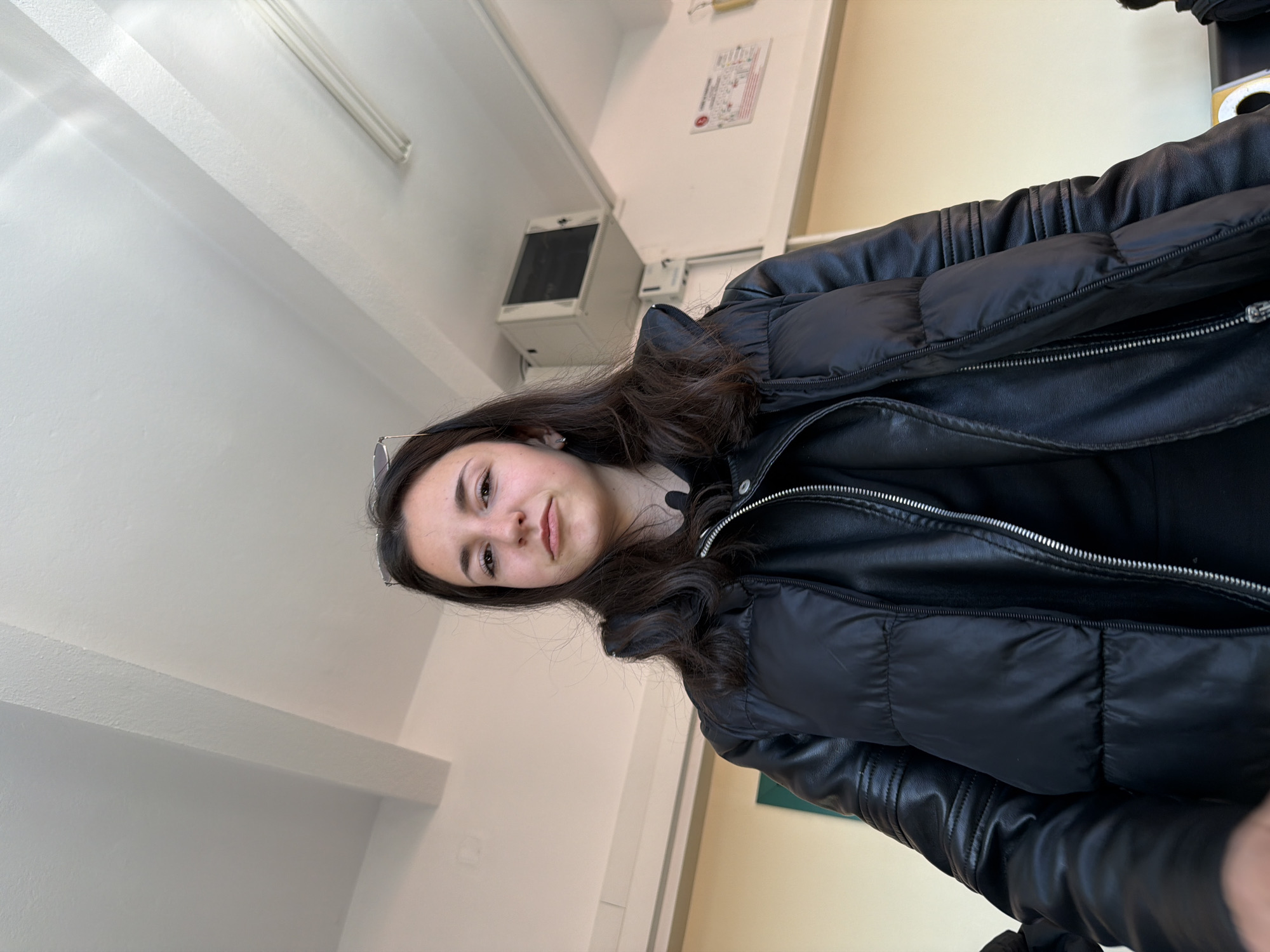 A young woman is indoors, likely in an institutional building, perhaps a school or university. She is wearing a black leather and puffy jacket. The room has white walls and a ceiling with exposed beams and light fixtures. There is an air conditioning unit mounted on the wall, and a bulletin board with notices. The photo was not taken in Panagyurishte, Bulgaria.