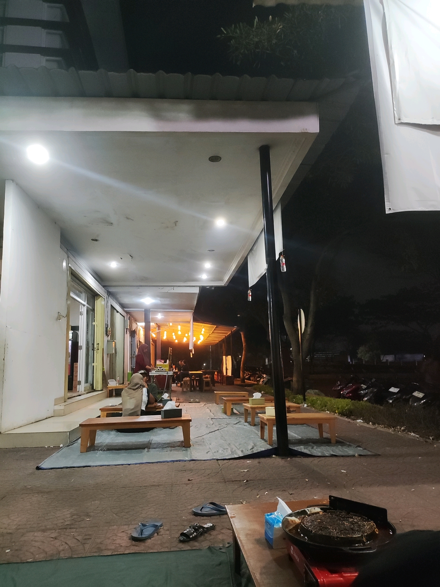 In Telagamurni, Indonesia, an outdoor seating area of a restaurant is depicted at night. A white building with an overhang forms the background. Seating arrangements include wooden tables and benches placed on gray tarps. People are seated at some of the tables, including a woman wearing a hijab. Strings of yellow lights hang overhead, illuminating the area. To the right, a row of motorcycles is parked along a grassy area. In the foreground, a portable stove with a cooking pan is set up on a table, with a tissue box and other items nearby. The ground is paved with reddish-brown tiles. Flip-flops are scattered on the ground. The scene suggests a casual dining or gathering atmosphere.