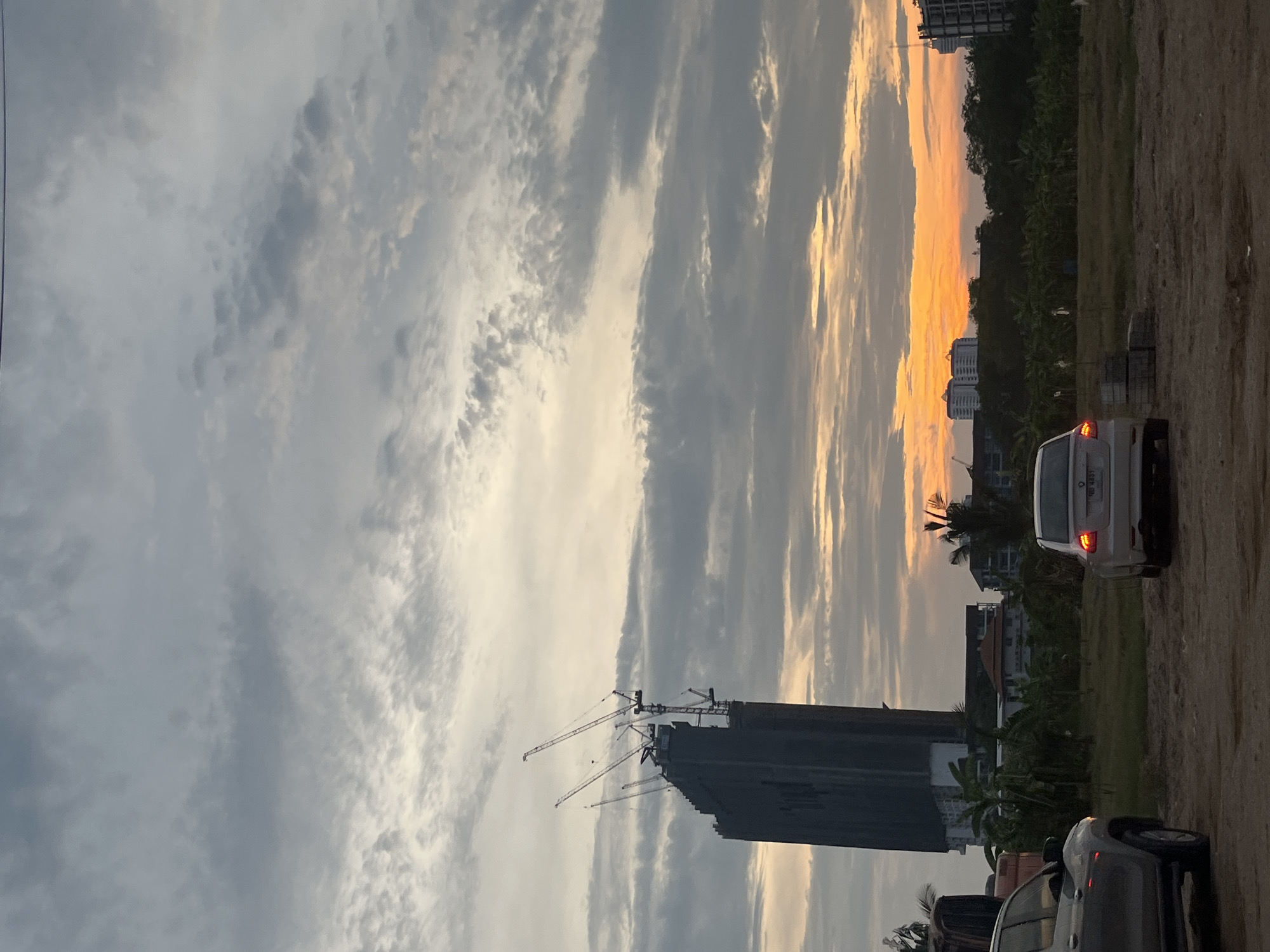 The image shows a cityscape at sunset in Pattaya City, Thailand. A tall building under construction dominates the view, with several cranes surrounding it. The sky is filled with clouds, some reflecting the orange and yellow hues of the setting sun. In the foreground, there is a dirt road with parked cars. A white sedan is visible with its taillights illuminated. Trees and other buildings are visible in the background.