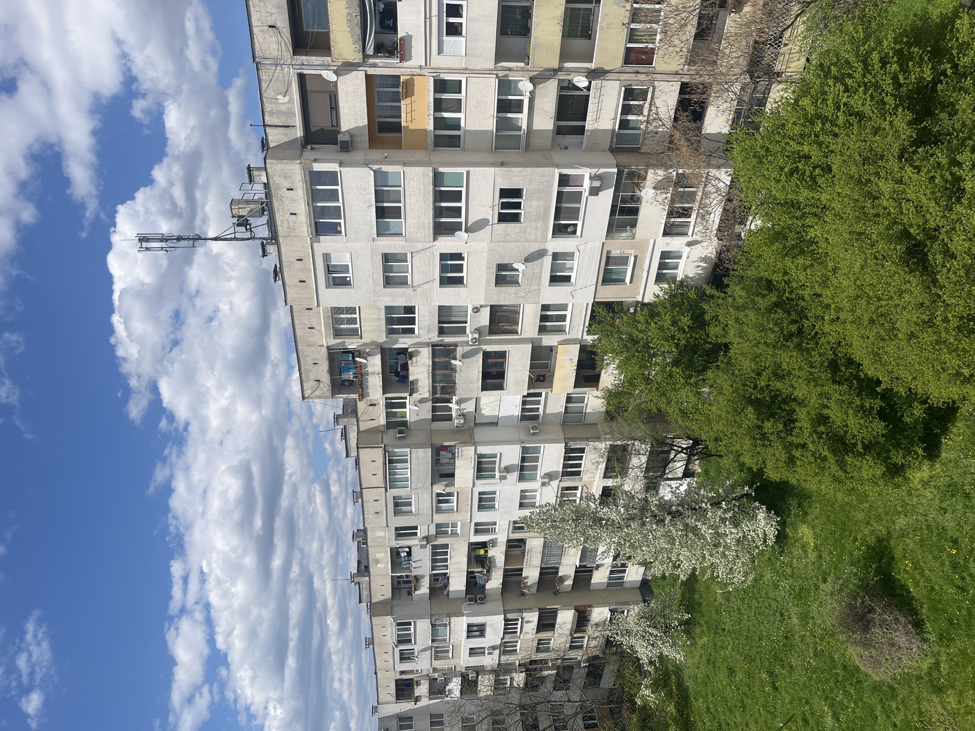 A daytime view captures a tall, multi-story apartment building typical of post-communist architecture, standing against a bright blue sky with scattered white cumulus clouds. The building, likely made of concrete or stucco, shows signs of age with varying shades of gray and some yellowed sections, particularly near balconies and window frames.
The facade is densely populated with numerous windows, many of which are modern white frames, while others appear older. Several small satellite dishes are mounted on the exterior walls, alongside various air conditioning units. Balconies extend from multiple apartments, some appearing enclosed or modified, others with laundry or personal items visible. A prominent, multi-component antenna structure is visible on the rooftop, suggesting communication equipment.
To the left of the building, a vibrant green landscape unfolds. Lush green trees with dense foliage occupy the mid-ground, with one striking tree covered in white blossoms, indicating spring or early summer. Below the trees, a patch of bright green grass is visible, featuring some small yellow wildflowers and a darker, smaller bush.
The scene depicts a typical urban residential environment in Sofia, Bulgaria, characterized by its functional architecture and surrounding greenery. No people or specific activities are clearly visible, and no discernible text is present on the building or in the environment. The weather appears fair and sunny, despite the presence of clouds.