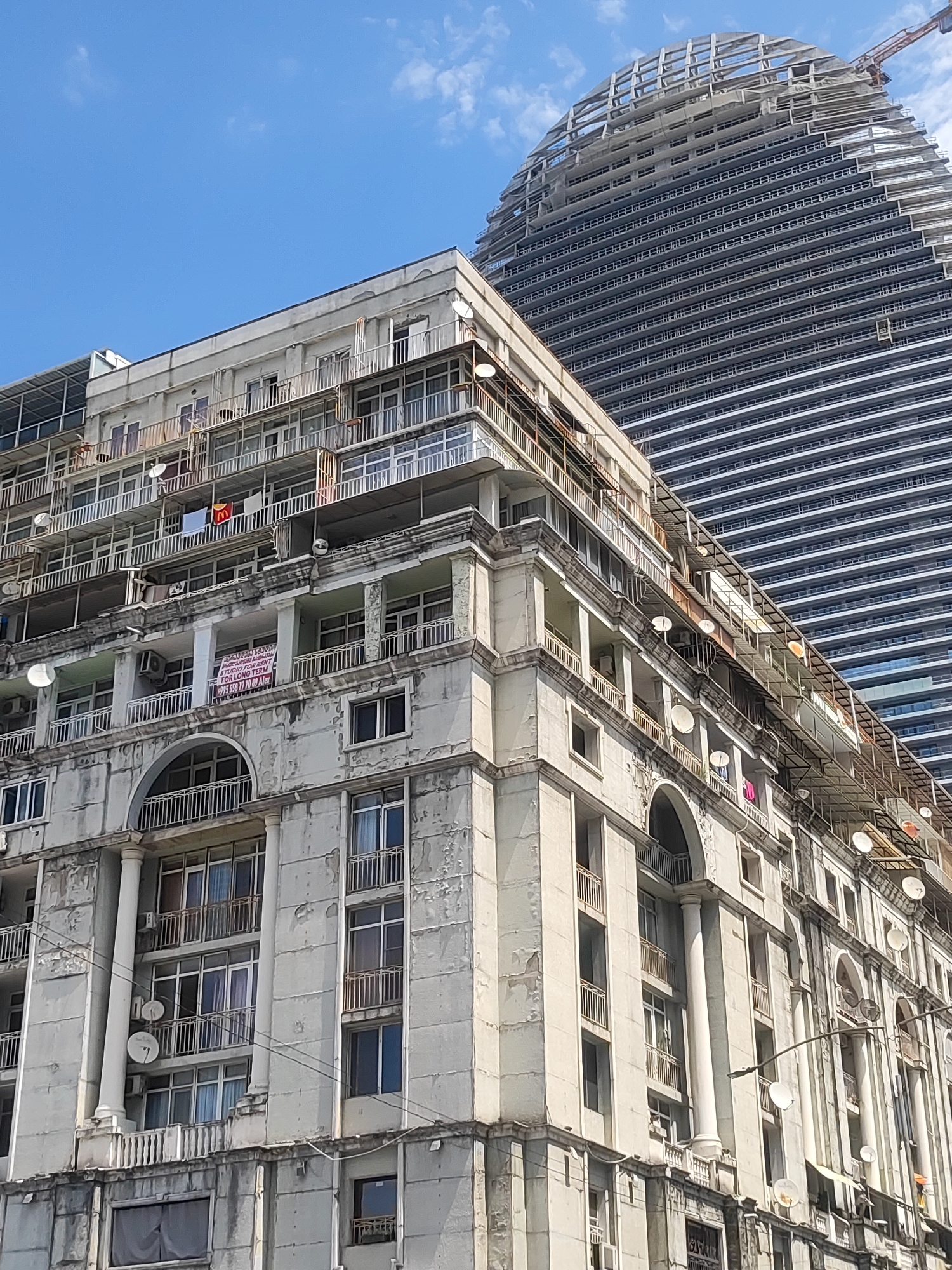 The image shows a wide-angle view of the city of Batumi, Georgia, on a sunny day. In the foreground, an old, weathered apartment building with multiple balconies and satellite dishes is visible. The building's facade is painted white, but it shows signs of age, with peeling paint and grime. Some balconies are enclosed with glass, while others have simple railings. Several windows are visible, with some curtains drawn. A banner with text in Georgian and English, advertising "STUDIO FOR RENT FOR LONG TERM", hangs from a balcony. On another balcony, a McDonald's flag is displayed. To the right and behind the old apartment building, a modern, curved skyscraper under construction dominates the background. The skyscraper is mostly concrete and steel, with rows of windows or balconies visible. A construction crane is positioned to the right of the skyscraper. The sky above is blue with scattered white clouds. The scene captures a contrast between older, established architecture and new development in the city.