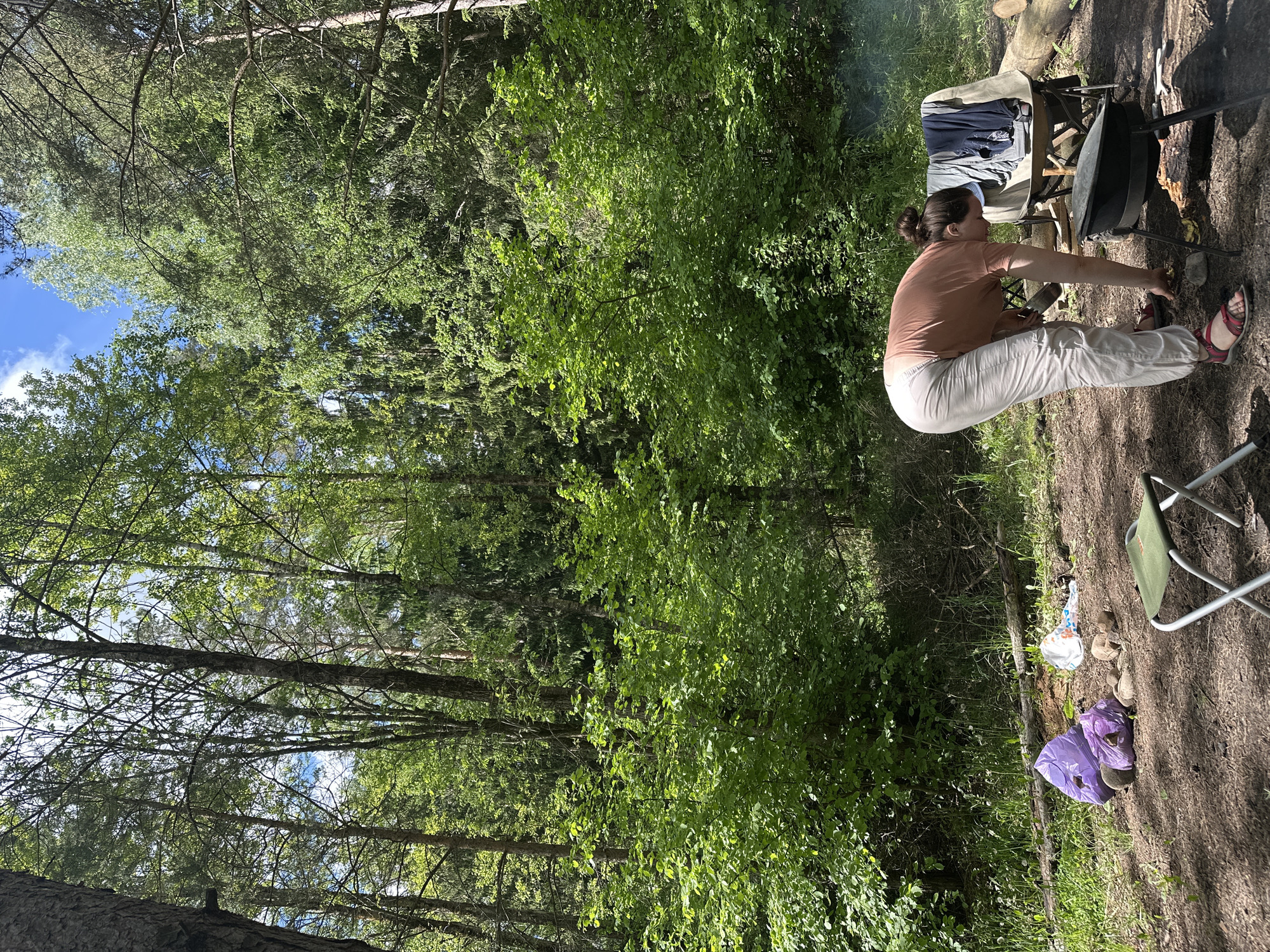 A woman is sitting next to a grill in a wooded area. She is wearing a pink shirt and white pants with red sandals. There is a lawn chair and clothing next to the grill. Another chair is to the right near a purple bag on the ground. The area is located in Юзуфоўскі сельскі Савет, Belarus.