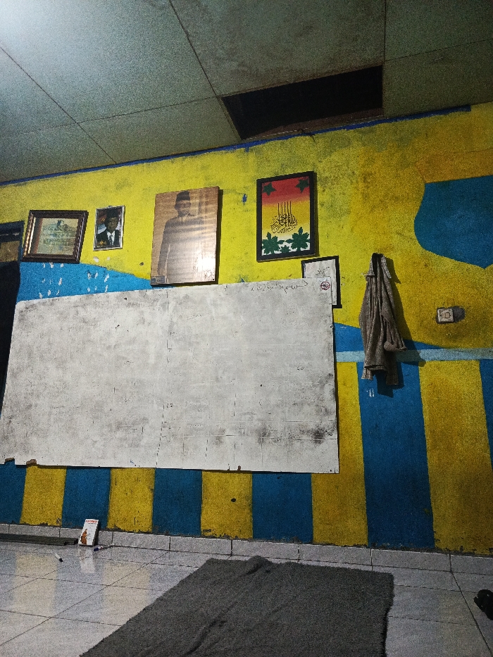 The image presents an interior view of a room, likely a classroom or communal space in Salatiga, Indonesia. The walls are distinctly painted in a vibrant, irregular pattern of yellow and blue. A large, worn white board with faint, illegible markings, possibly including Arabic script and numbers, is prominently centered on the wall. To the right of the whiteboard, a smaller framed document with indistinct text is visible.
Above the whiteboard, a collection of framed items hangs on the yellow wall. From left to right, there is a small framed document, followed by a framed portrait of a man in a suit. The most prominent portrait is a larger, rectangular one depicting a man wearing a peci (traditional cap) and a suit, strongly suggesting an influential Indonesian figure such as Sukarno. Further right, a framed artwork displays intricate Arabic or Jawi calligraphy in black script, set against a red and orange gradient background and adorned with green, leaf-like designs.
To the far right, a light-colored cloth or garment hangs from a hook on the blue section of the wall, below which is a white electrical outlet or light switch. The ceiling features square, textured panels, with a noticeable dark, rectangular opening or missing panel in the upper section. The floor is covered with light-colored square tiles, upon which a dark grey, rectangular rug or mat is placed. Near the bottom left, a small book or box and a piece of white packaging or paper are visible on the tiled floor.
The lighting is artificial and somewhat dim, suggesting the scene takes place indoors, possibly in the evening or at night. No direct activity is depicted, making the scene static. The visible portraits and calligraphy are significant cultural cues for Indonesia.