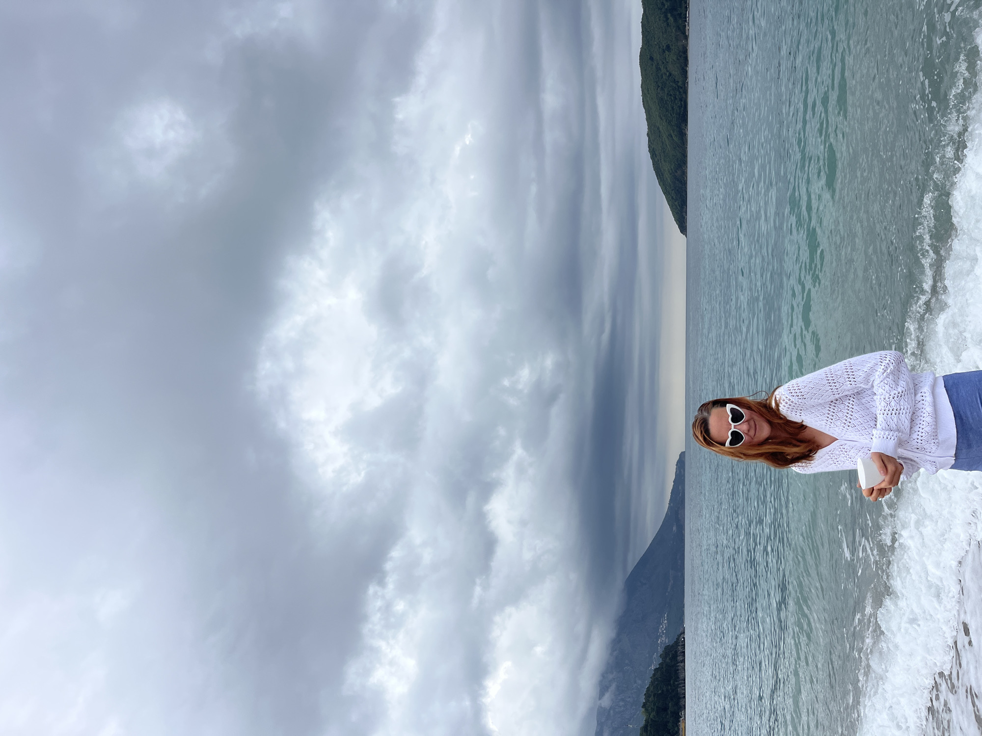 The image, rotated 90 degrees clockwise from its intended orientation, captures a young woman posing on a beach. She stands near the water's edge, where gentle waves break with white foam around her.
The woman has long, reddish-brown hair and is smiling warmly at the camera. She is wearing a white, open-knit cardigan over a lighter top, light blue trousers or jeans, and distinctive white, heart-shaped sunglasses. In her hands, she holds a small, white, rectangular object, possibly a phone or a smooth pebble.
Behind her, the calm, blue-green sea extends towards the horizon. A large, dark green mountain, covered in dense foliage, rises majestically and slopes down into the water on the right side of the frame (when viewed correctly). Another, hazier mountain range or distant landmass is visible further in the background to the left. The sky is heavily overcast with thick, grey clouds, indicating a cloudy or possibly stormy day. The serene setting, with mountains meeting the sea, is characteristic of Budva, Montenegro, where the image was taken. There is no visible text in the image.