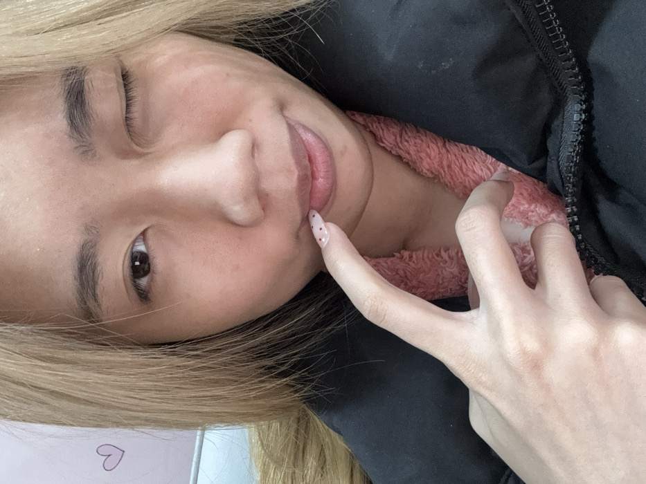A close-up photograph depicts an individual with light blonde hair, winking their proper right eye while their proper left eye remains open. The subject's proper right index finger, which features a clear or light-pink nail with small reddish-brown circular embellishments, is positioned against their slightly parted proper lower lip. The individual is dressed in a black puffer jacket layered over a pink, textured fleece-like garment. The background consists of a blurred, light-colored surface displaying a faint purple heart graphic on the proper left and a light blue linear element on the proper left. The setting is an indoor environment located in Shapingba District, China.