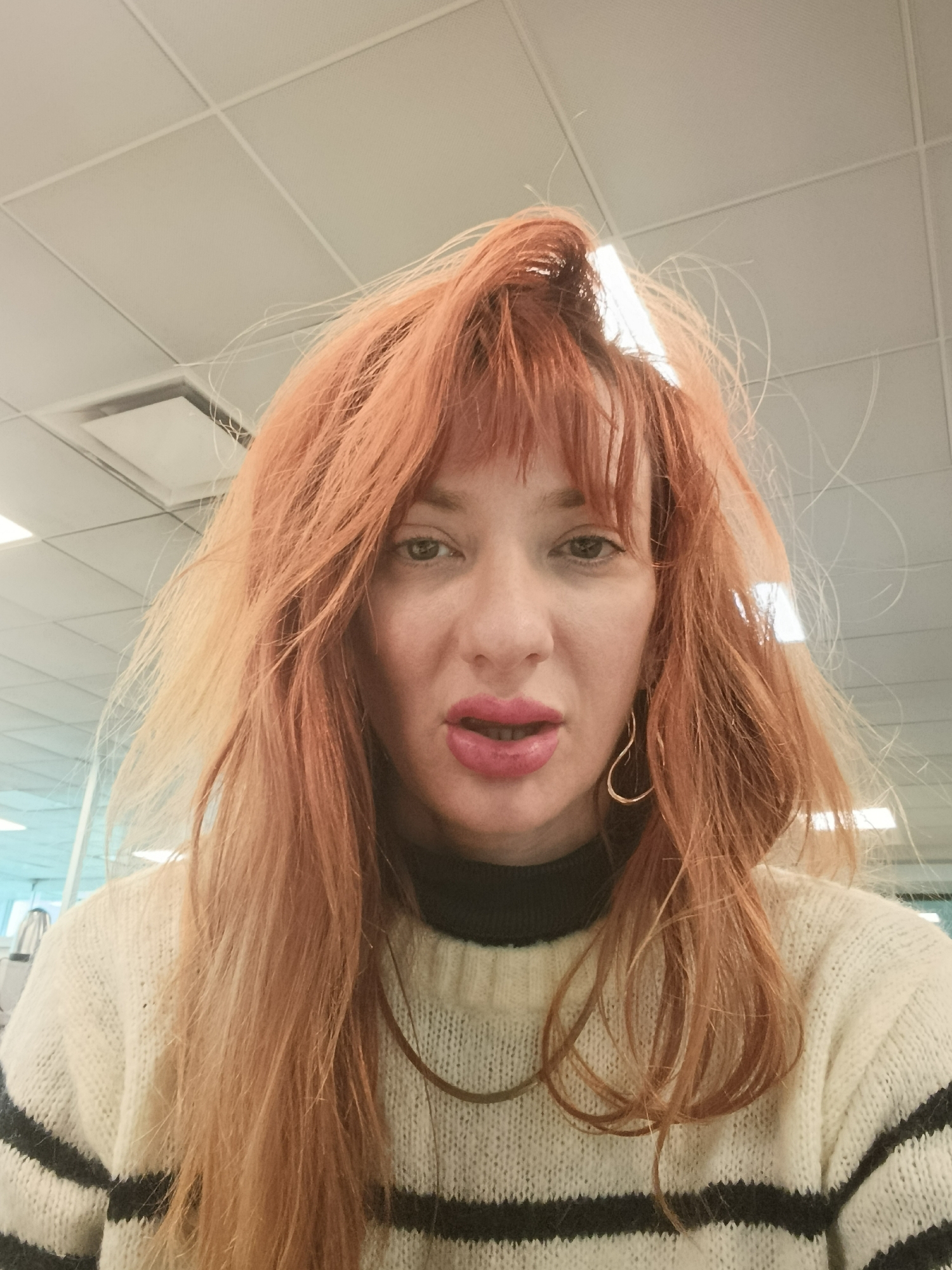 A woman with long, orange-red hair is facing the camera indoors. She wears a cream-colored sweater with black stripes and a black turtleneck underneath. She has gold hoop earrings and a gold necklace. Her lips are painted pink. The background is a white ceiling with fluorescent light fixtures. The location in the city of Buenos Aires, Argentina is not discernible from the image.