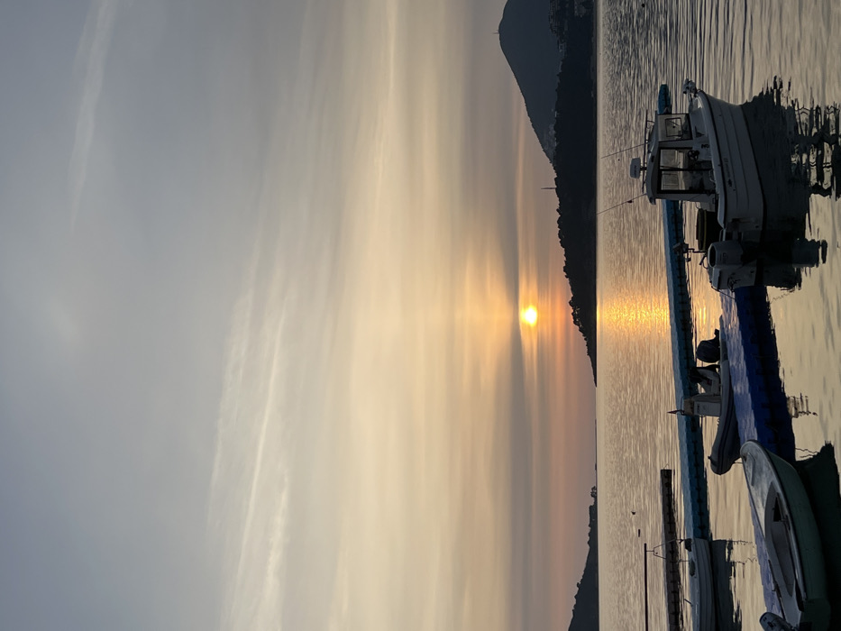 The image depicts a sunset over a calm body of water in Budva, Montenegro. The sun, a bright orange orb, is positioned low on the horizon, slightly left of the image's center, casting a prominent reflection path of orange light across the water. In the foreground, several vessels are moored to floating pontoons. On the far left, a light green dinghy is partially visible, connected to a lighter-colored floating structure. Centrally, a white rigid-hull inflatable boat (RIB) equipped with an outboard motor is secured to a blue modular floating pontoon. To its right, a larger white cabin cruiser, featuring two outboard motors, a radar dome, and visible fishing rods, is also docked at the same blue pontoon. Dark, tree-covered landforms frame the horizon: a smaller peninsula or island is present on the left, and a larger, mountainous peninsula is visible on the right, distinguished by a communication tower near its summit. The sky transitions from a vibrant orange and yellow hue near the horizon to a pale blue-grey towards the top, with faint, diffused cloud formations. No individuals or explicit human activities are discernible within the frame.
