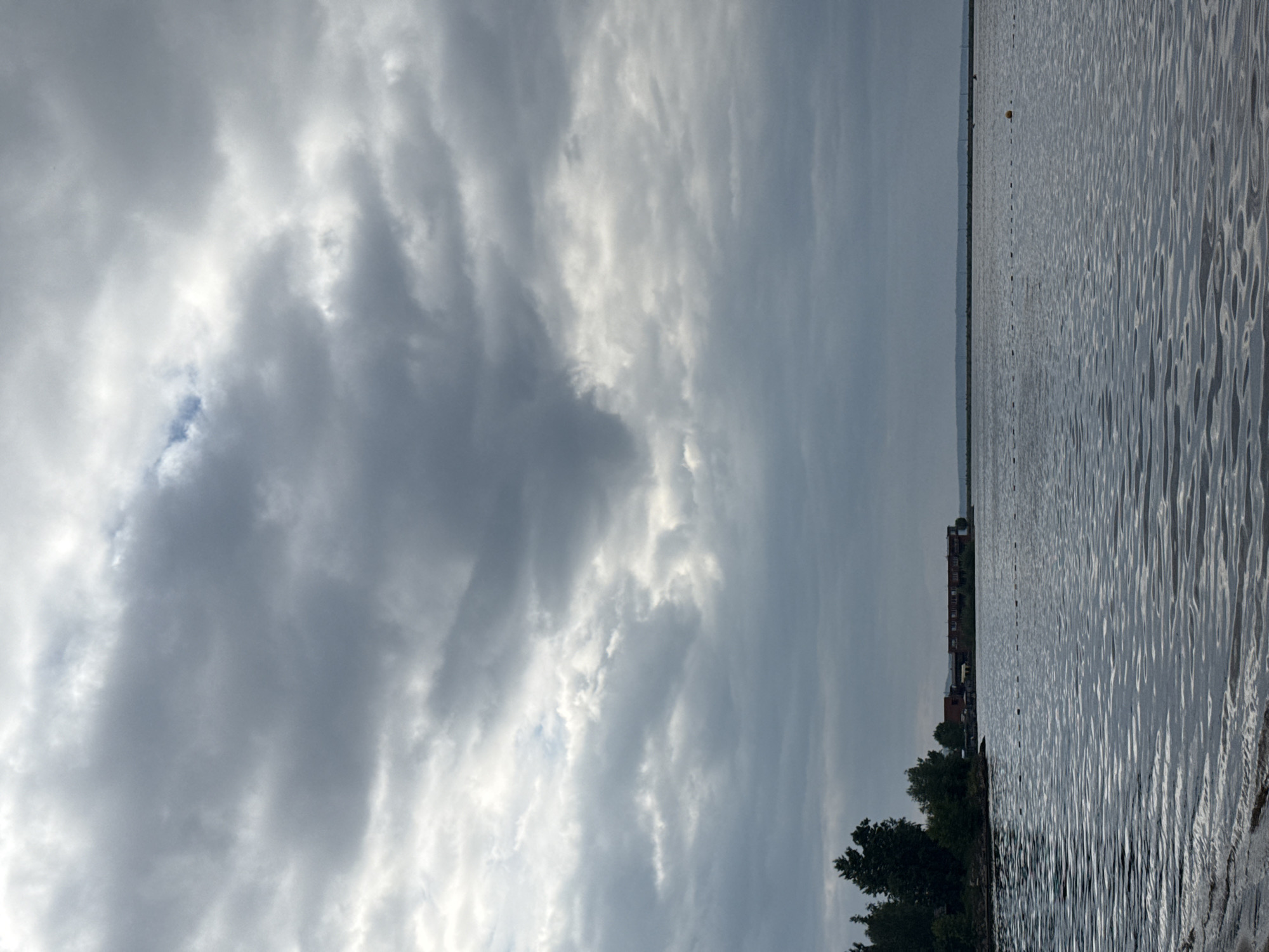 The image depicts a waterfront scene under a cloudy sky. In the foreground, the calm water of a large body of water, likely the Kama River, laps at a sandy shore. The water's surface is rippled, reflecting the overcast sky. In the midground, a line of buoys stretches across the water. To the left, a wooded shoreline with trees is visible, and beyond that, a long, multi-story building with a red roof is situated along the water's edge. The building appears to be part of a residential or recreational complex. The background is dominated by a dense layer of dark gray clouds that cover the entire sky. The overall impression is one of a peaceful but somber day, possibly in the city of Chaikovsky, Russia, given its prominent location on the Kama River.