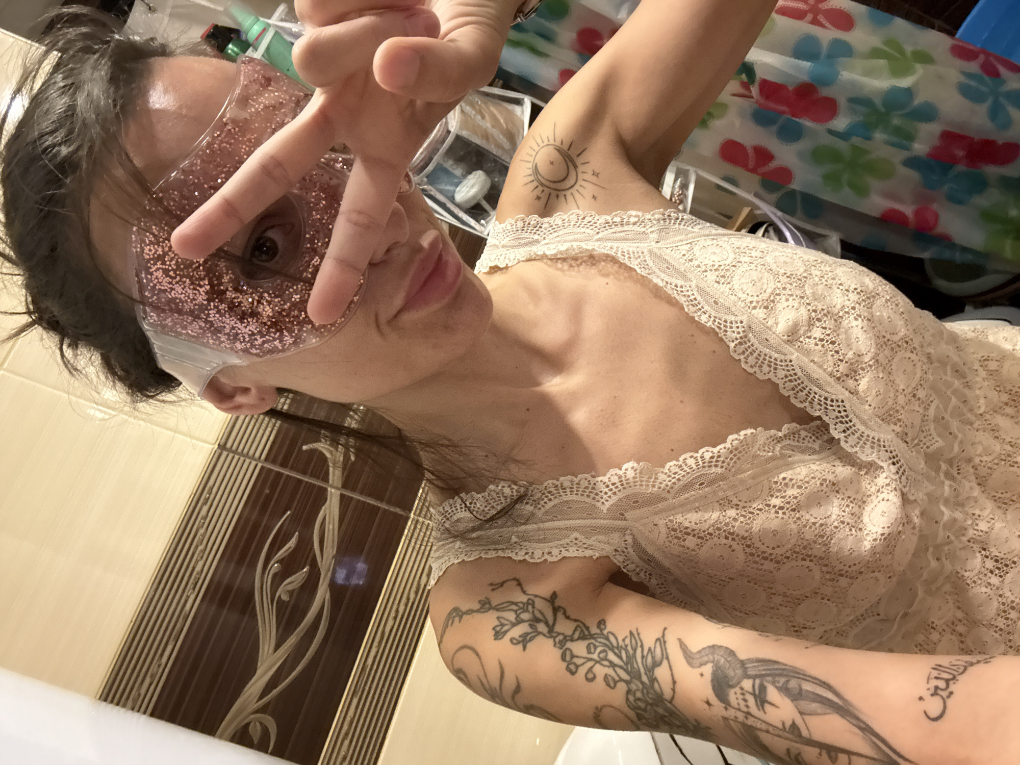 A person, likely female, is featured in a close-up selfie, taken from a low angle in what appears to be a bathroom in Pleven, Bulgaria. She has dark hair and is wearing a light-colored, possibly off-white, lace top.
Her face is partially covered by a translucent pink gel eye mask filled with glitter, designed for relaxation or cooling. She holds her right hand in front of her face, displaying a 'peace sign' with two fingers extended over the eye mask. Her lips are slightly pursed, conveying a playful or confident expression.
Several tattoos are visible on her arms. On her right upper arm, there is a distinct tattoo of a sun with a crescent moon inside, surrounded by radiating lines. On her left forearm, a more intricate tattoo features flowing botanical elements, possibly incorporating a bird with horns or antlers. Below this, legible Arabic script is tattooed, reading "والتين" (wal-teen), meaning "and the fig."
The background indicates an indoor setting. A colorful shower curtain or textile with red, blue, and green floral patterns is visible. A tiled wall, featuring vertical brown and beige stripes interspersed with a lighter, decorative floral motif, is also present. A white surface, possibly the rim of a bathtub or sink, is seen in the foreground. The overall atmosphere of the image is casual and relaxed, capturing a personal moment of self-care or playful interaction with the camera.