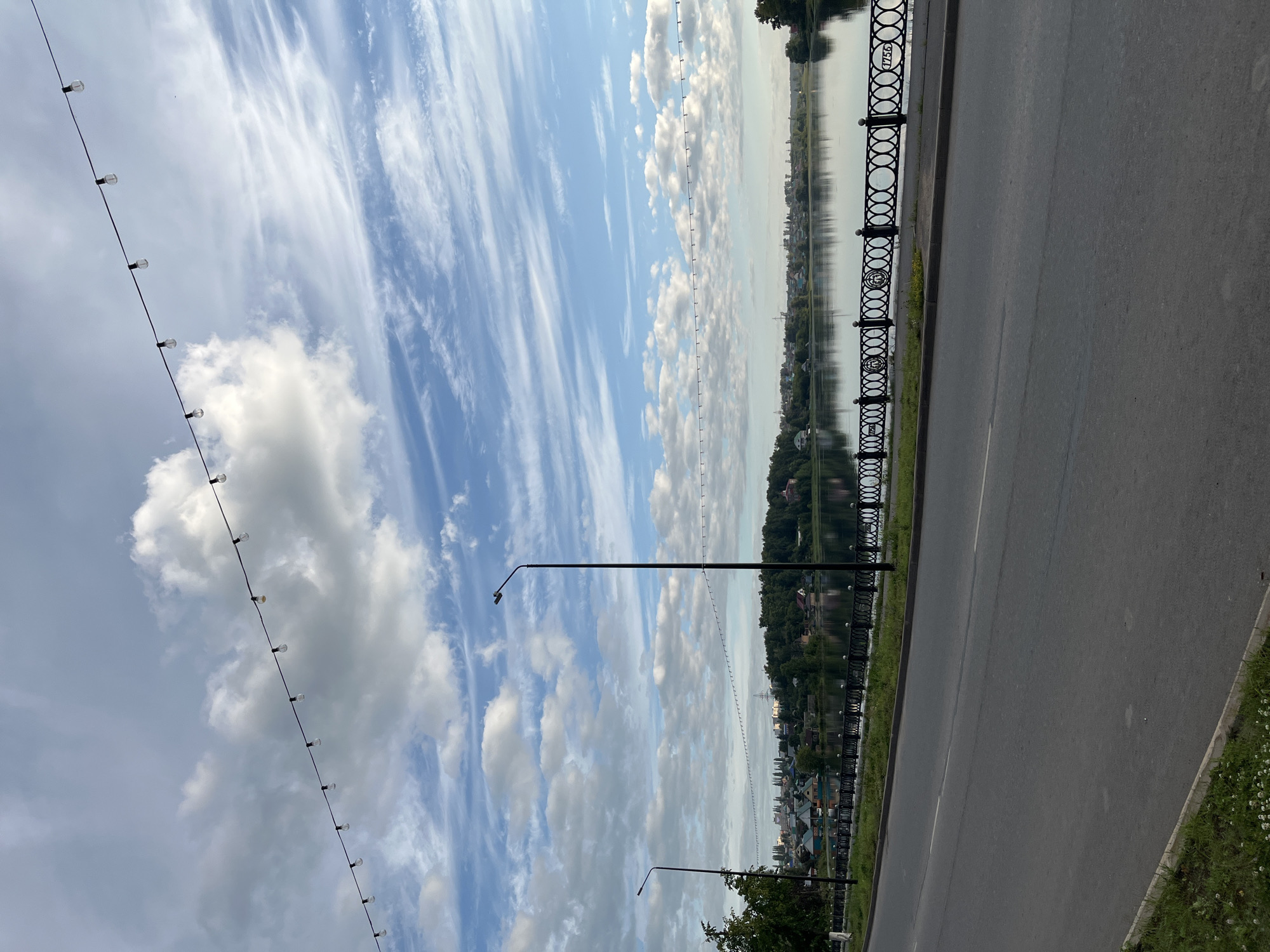 The image shows a scenic view of the Amur River in Blagoveshchensk, Russia. The riverfront promenade is lined with a decorative black metal fence, featuring circular patterns and the year "1755" embossed on some sections. Beyond the fence, the calm waters of the Amur River reflect the sky and the lush green trees and buildings on the opposite bank. A paved road runs alongside the river, with a grassy verge and a few small white flowers at the edge. Two streetlights are visible, one tall and one shorter, spaced along the promenade. Above, a string of lights with visible bulbs is strung across the sky. The sky is partly cloudy with streaks of cirrus clouds, suggesting it is either early morning or late afternoon. There are no people or animals visible in the image.