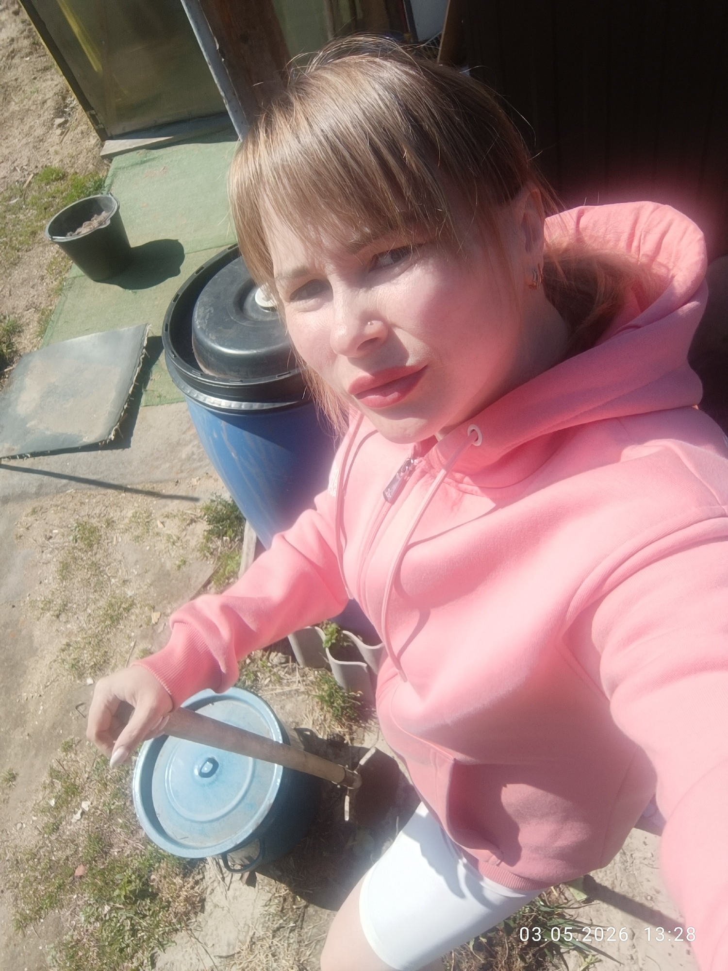 A woman is holding a wooden stirring stick over a blue-lidded pot, possibly outdoors in Chaikovsky, Russia. She is wearing a pink hoodie, white leggings, and has light brown hair with bangs. A blue barrel and black bucket are visible in the background, along with other gardening items and sparse grass. The date "03.05.2026" is displayed at the bottom right corner of the image.