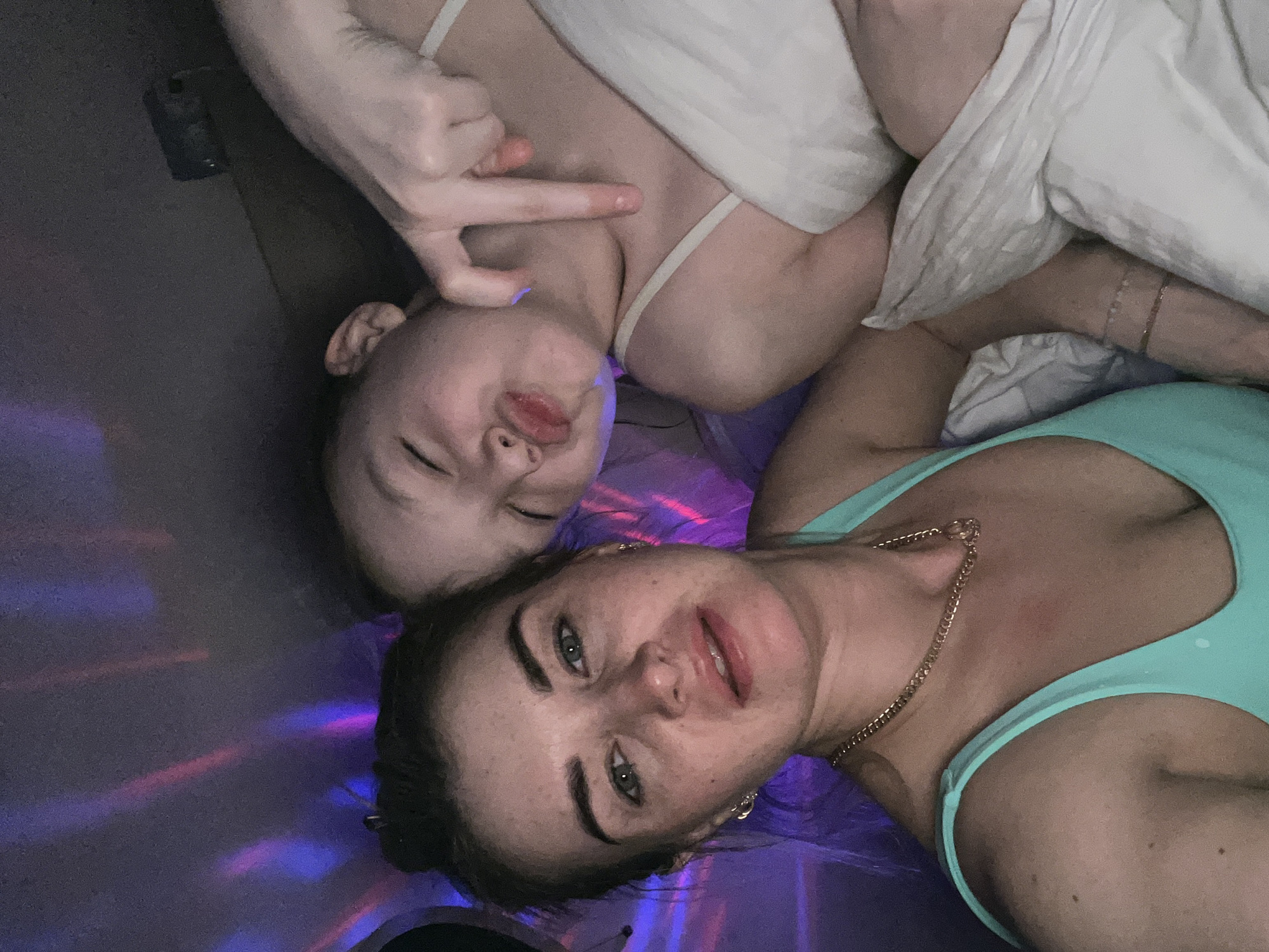 Two individuals are lying down, facing the camera. One person, likely a child, is wearing a white top and has their eyes closed, with a hand making a gesture towards their face. The other person is wearing a teal top and a gold necklace. The background is illuminated with purple and pink lights. The location is unspecified beyond the image's content. There is no indication of the image's location being in the city of Chaikovsky, Russia.