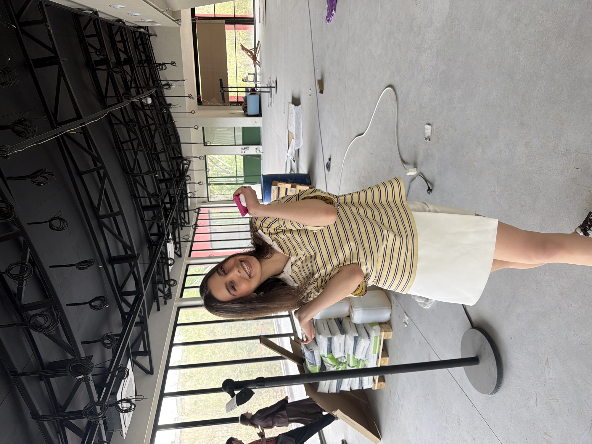A woman stands in a large, unfinished interior space. She is wearing a yellow and white striped shirt and a white skirt. She is holding a pink object, possibly a phone. In the background, there is a metal grid-like structure on the ceiling, along with large windows that reveal trees outside. Bags of what appears to be construction material are stacked on a wooden pallet. A black metal pole with a circular base is prominent in the foreground. Other people are partially visible in the background. The location is likely an industrial or commercial space undergoing construction. I am unable to identify the location as Yerevan, Armenia.