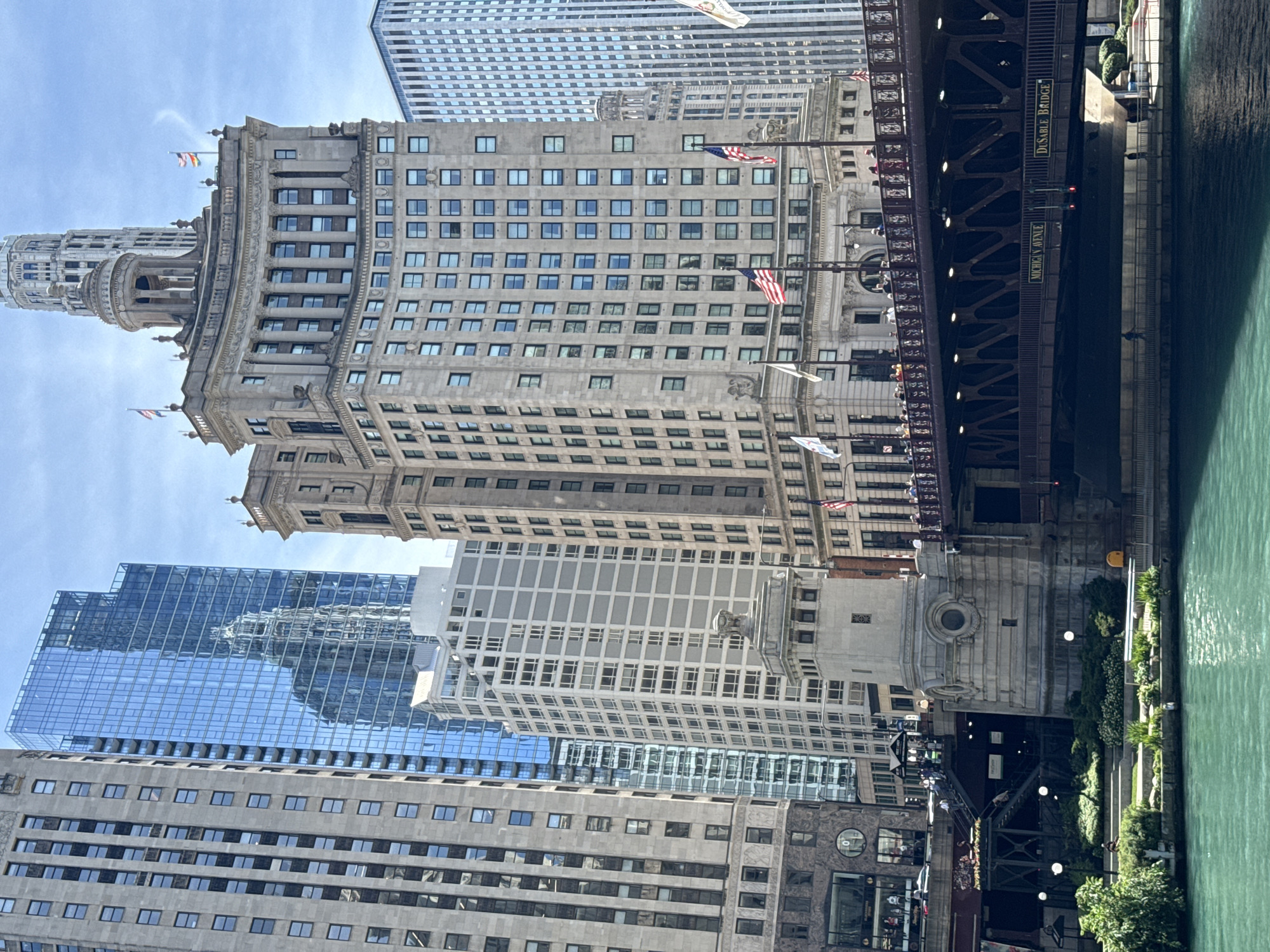This image captures a scenic view of Chicago, USA, featuring a collection of imposing skyscrapers and a bridge spanning a body of water. The scene is bathed in bright daylight under a clear sky, suggesting a pleasant weather day.
The central focus is a large, ornate stone building with numerous windows, characteristic of classic Chicago architecture. To its left, a taller, more modern skyscraper with a reflective glass facade stands out, its surface mirroring the surrounding buildings and sky. Another, more distant skyscraper with a distinctive pattern of horizontal lines is visible behind these structures.
A prominent bridge, likely a bascule bridge, crosses the water on the right side of the image. The bridge is a dark metal structure with visible supports and railings. Signage on the bridge reads "MICHIGAN AVENUE" and "DU SABLE BRIDGE," identifying its location and name. The water, a vibrant turquoise, flows beneath the bridge, its surface rippling gently.
Along the riverbank, a pedestrian walkway is lined with lush greenery and trees, with several lampposts illuminating the path. People can be seen walking and standing on the bridge and along the riverfront, suggesting the area is a popular public space. A few American flags are displayed on poles, adding a patriotic touch to the urban landscape.
The overall impression is one of a bustling city scene, showcasing the blend of historical and contemporary architecture that defines Chicago's skyline, with the river and bridge adding a dynamic element to the composition.
