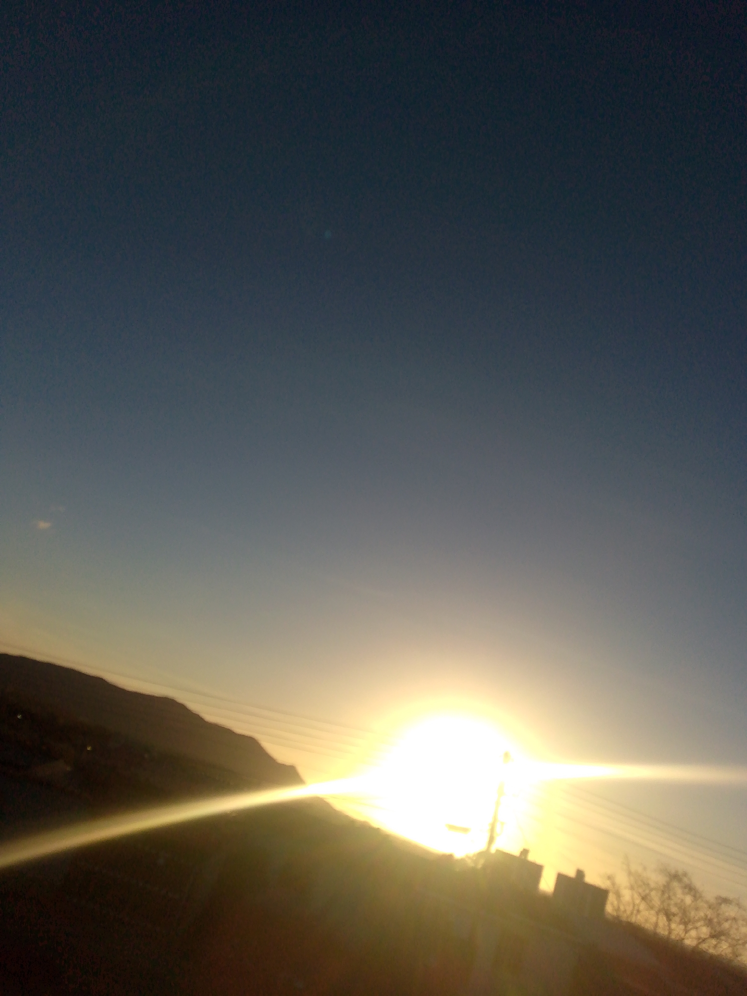 The image depicts a sunrise or sunset, potentially in Juan Griego, Venezuela. The bright sun is low on the horizon, obscured partially by a silhouette of a hill or mountain. In the foreground, there appear to be buildings and vegetation, also in silhouette against the intense sunlight. The sky is transitioning from a darker shade at the top to a lighter color near the horizon, indicating the presence of the sun. Power lines are visible.