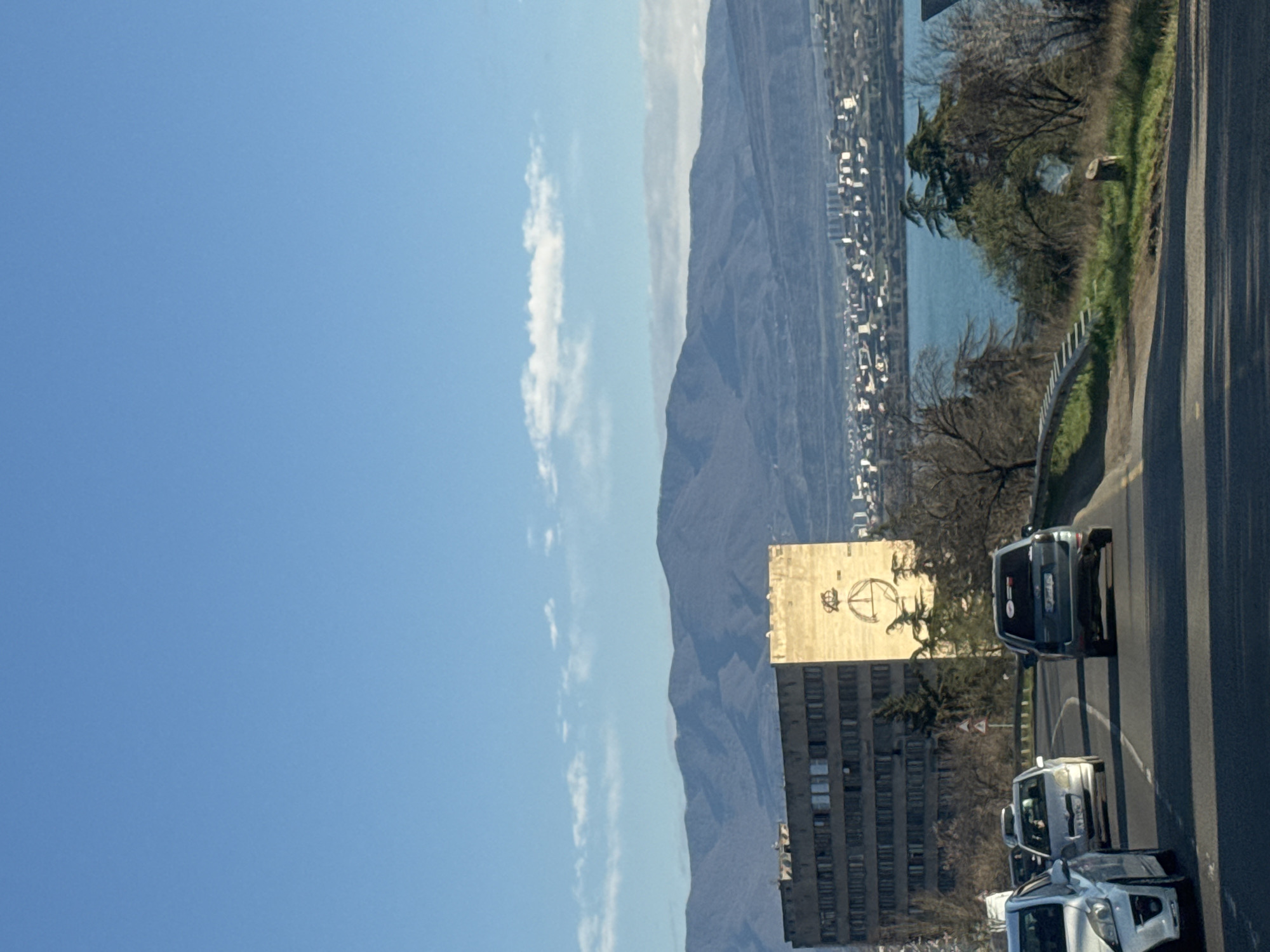 The image depicts a street scene in Tbilisi, Georgia. A multi-lane road is visible with at least three vehicles traveling on it. A large, Soviet-era building stands adjacent to the road, featuring a faded emblem on its facade. In the background, a mountain range rises towards a blue sky with scattered clouds. Between the buildings and the mountains, a body of water and a densely populated urban area can be seen. The scene is bathed in sunlight, suggesting a clear day.