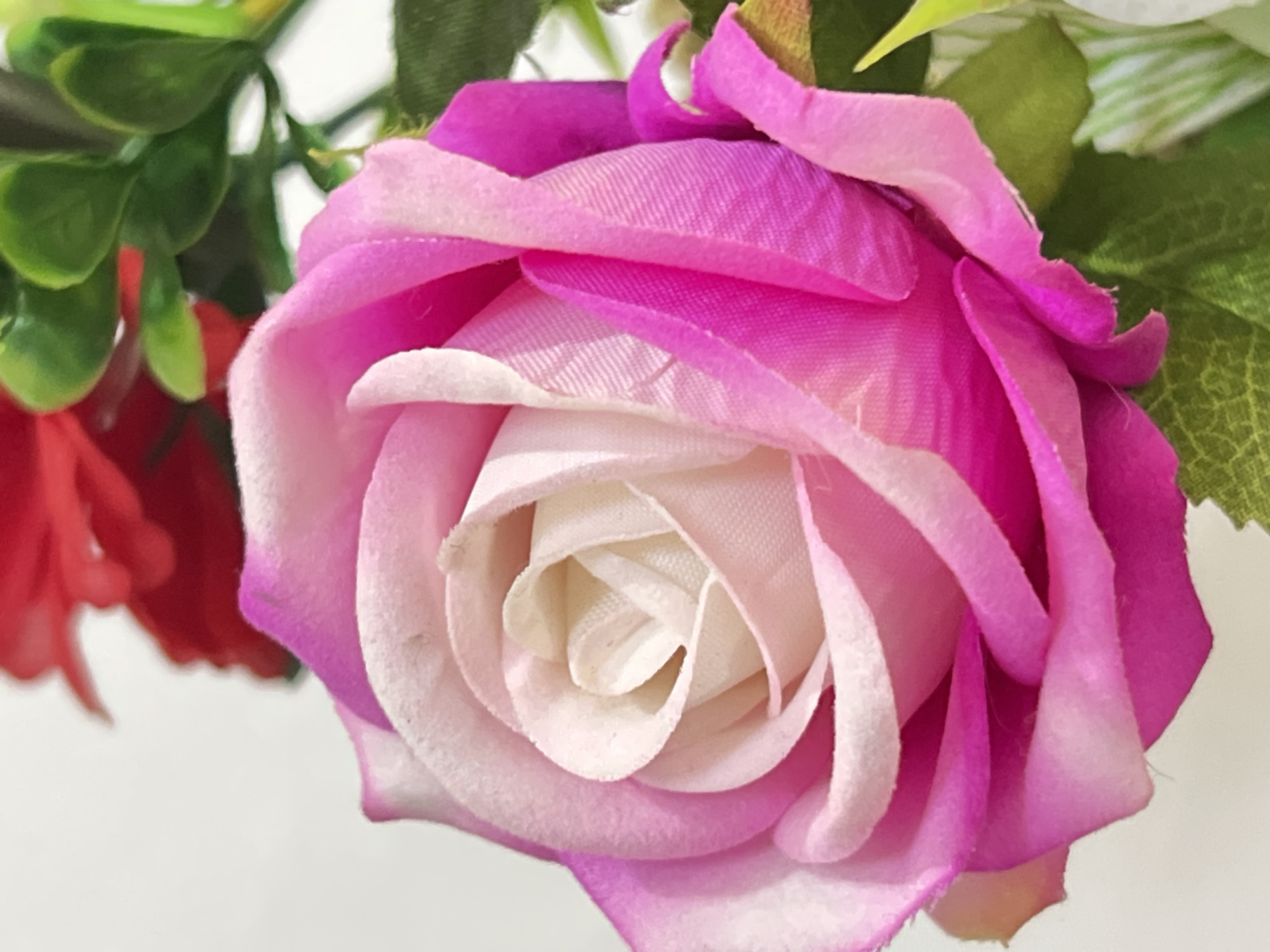 A close-up, full-frame shot focuses on a vibrant, artificial pink rose with a cream-colored center. The rose is the dominant subject, with its soft, fabric petals arranged in a spiral. The petals transition from a pale cream at the core to shades of light pink and then a deep fuchsia at the edges. Sunlight subtly illuminates the petals, highlighting their texture and layered structure. In the background, out of focus, are hints of red flowers and green foliage, suggesting a bouquet or arrangement. The lighting is soft and diffused, creating a gentle glow on the rose. The image is devoid of human presence or activity, presenting a static and aesthetic composition.