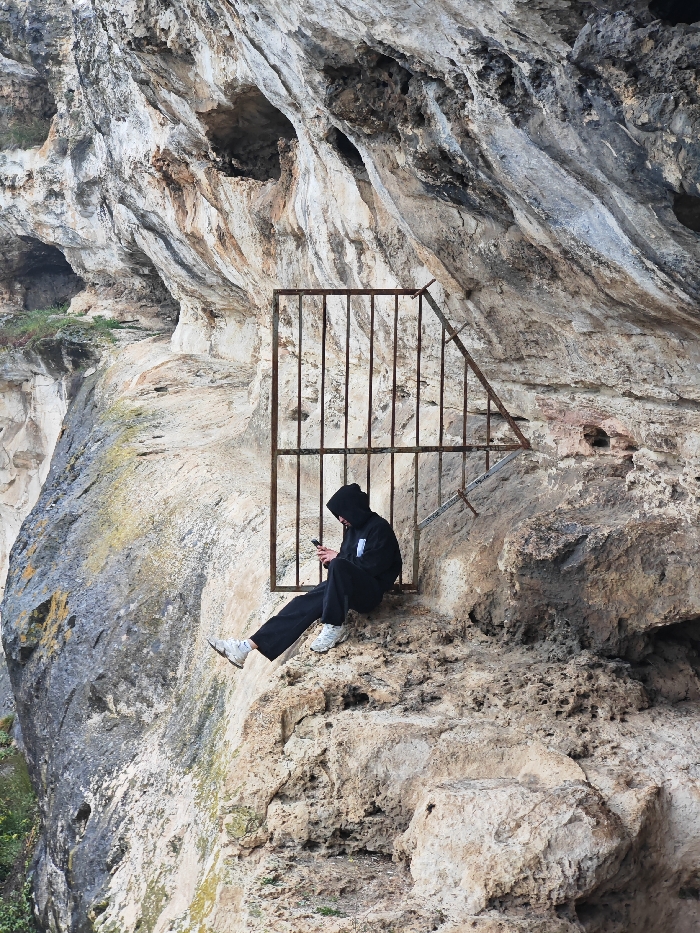 A single individual is observed sitting on a natural rock ledge against a prominent limestone cliff face near Shumen, Bulgaria. The person is dressed in a black hooded sweatshirt with the hood drawn, obscuring the upper face, black trousers, and white athletic shoes. They are focused on a dark-colored mobile phone, held in both hands, with their gaze directed downwards towards the screen. Immediately behind and to the right of the individual, a rusted metal gate, constructed from vertical and diagonal bars, is fixed into the rock, partially obstructing a cave opening. The cliff face exhibits varied textures and colorations, predominantly in shades of white, gray, and beige, with scattered patches of sparse green vegetation.