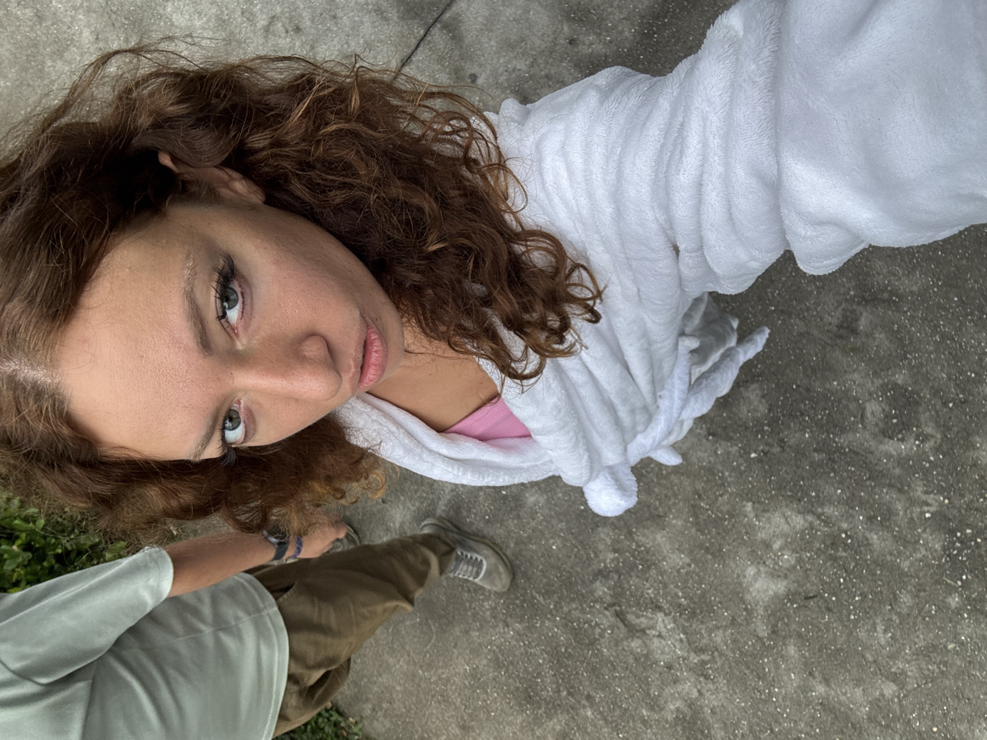 A selfie shot shows a fair-skinned woman with curly brown hair and light-colored eyes. She is wearing a white terry cloth robe over what appears to be a pink tank top. To her left, a person is partially visible, wearing a light green shirt and brown pants, along with gray shoes. The background consists of a gray concrete surface, possibly a sidewalk, and some green foliage in the corner. The image's composition is angled, creating a distorted perspective of the scene.