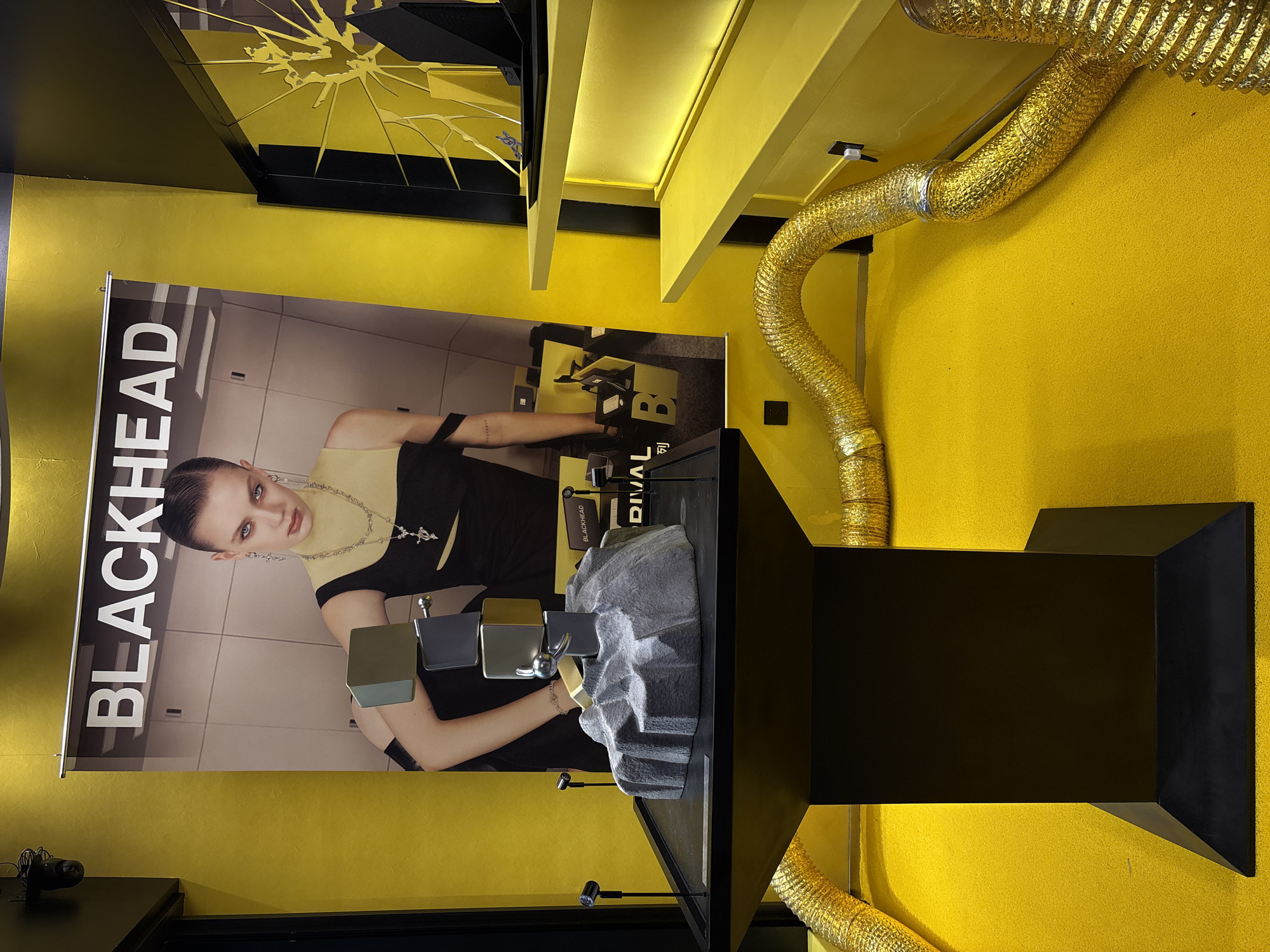 The image shows an indoor retail display, likely a jewelry store, with a predominantly yellow and black color scheme. A large banner featuring the word "BLACKHEAD" and a model wearing jewelry hangs on the wall. In the foreground, a black pedestal supports a grey, rock-like sculpture with several stacked metallic cubes and a sphere. Two small spotlights are positioned to illuminate the display. Flexible, gold-colored tubes are draped across the floor and up the sides of yellow shelves. In the background, a yellow wall and yellow shelves are visible, with a black display unit that has a cracked, reflective surface. The overall environment appears to be a commercial space designed to showcase products. The specific location is in the Changning District, China.