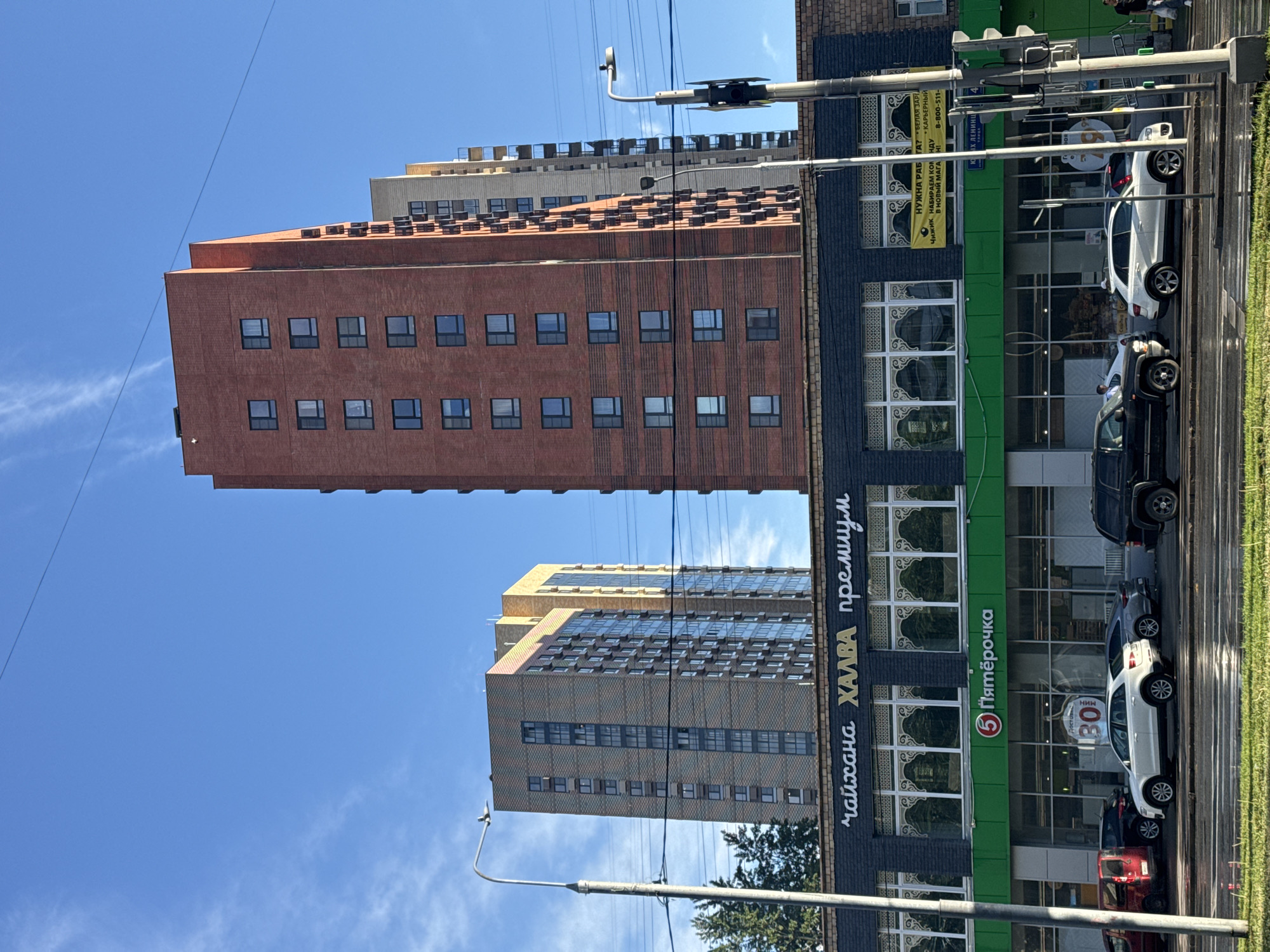 The image depicts a street scene in Moscow, Russia, featuring two tall residential buildings in the background and a storefront in the foreground. The taller building is brick-colored with rows of square windows, and the other is a light brown. The sky is clear and blue. The storefront has "Пятёрочка" (Pyaterochka) grocery store logo, a chain of stores. It also has signs written in Cyrillic script. Several parked cars line the street in front of the building. Overhead power lines are visible crossing the scene.