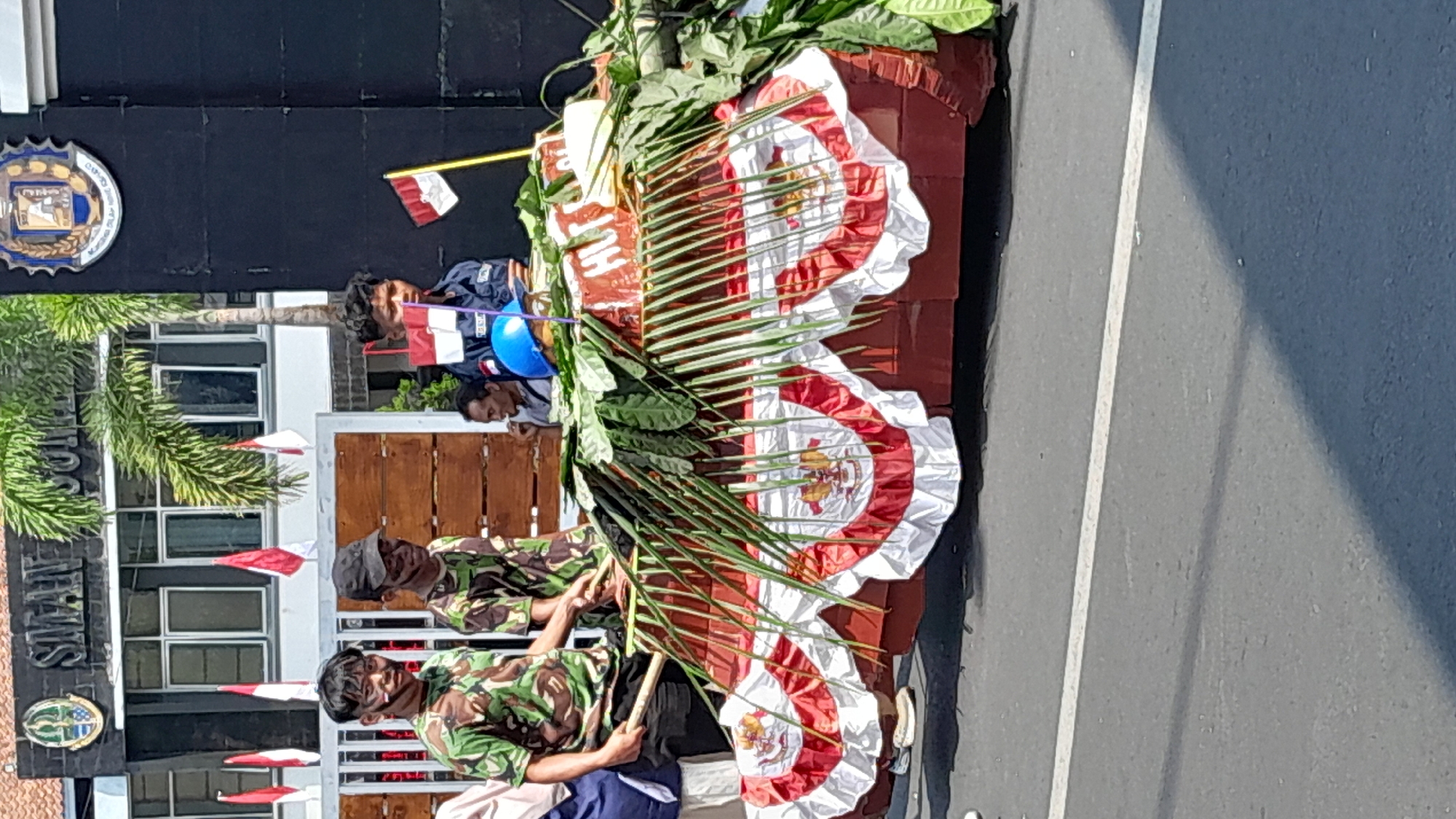 In Sumedang, Indonesia, in front of the SMA NEGERI 1 SUMEDANG building, a parade float is being displayed. Three young men, two in camouflage uniforms with face paint, stand by the float. The float is decorated with red and white bunting featuring the Indonesian coat of arms, and adorned with green leaves and palm fronds. A blue hard hat and a small Indonesian flag are visible on top. Other individuals are also present, some observing the float. The scene appears to be a public event, possibly a celebration or commemoration.